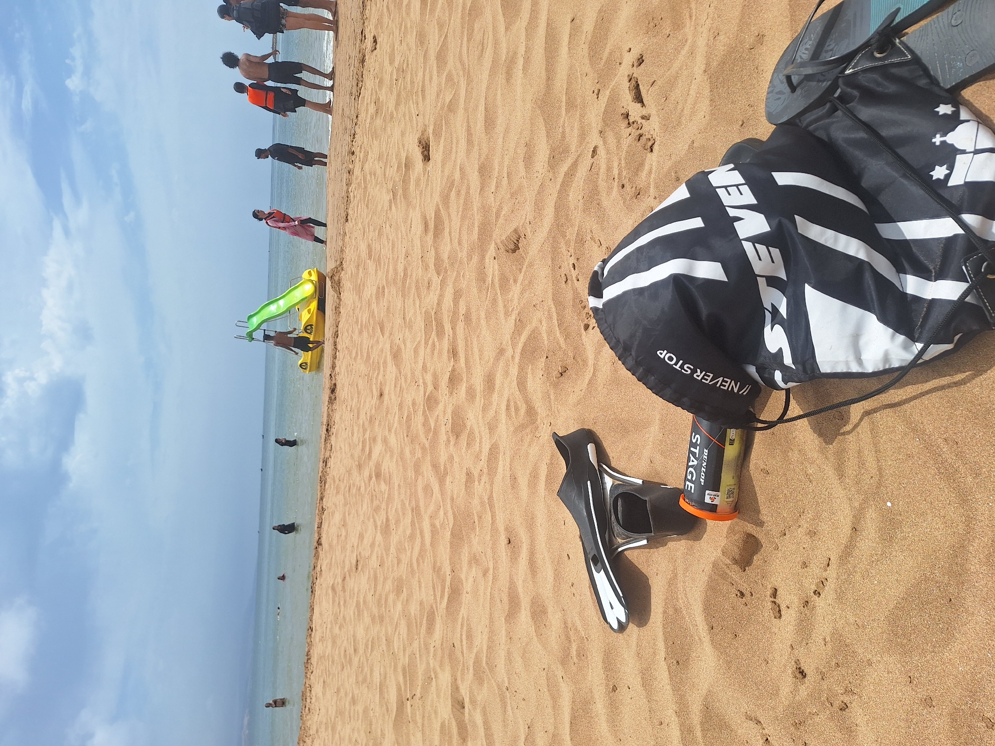 The image depicts a sandy beach in Imsouane, Morocco, with the ocean in the background. In the foreground, on the sand, are a pair of black and white fins, a tube of Dunlop Stage tennis balls, and a black drawstring bag with white stripes and logos. Further up the beach, several people are visible, some wading in the water and others standing on the shore. A bright yellow pedal boat with a green slide is also present in the water. The sky is partly cloudy.