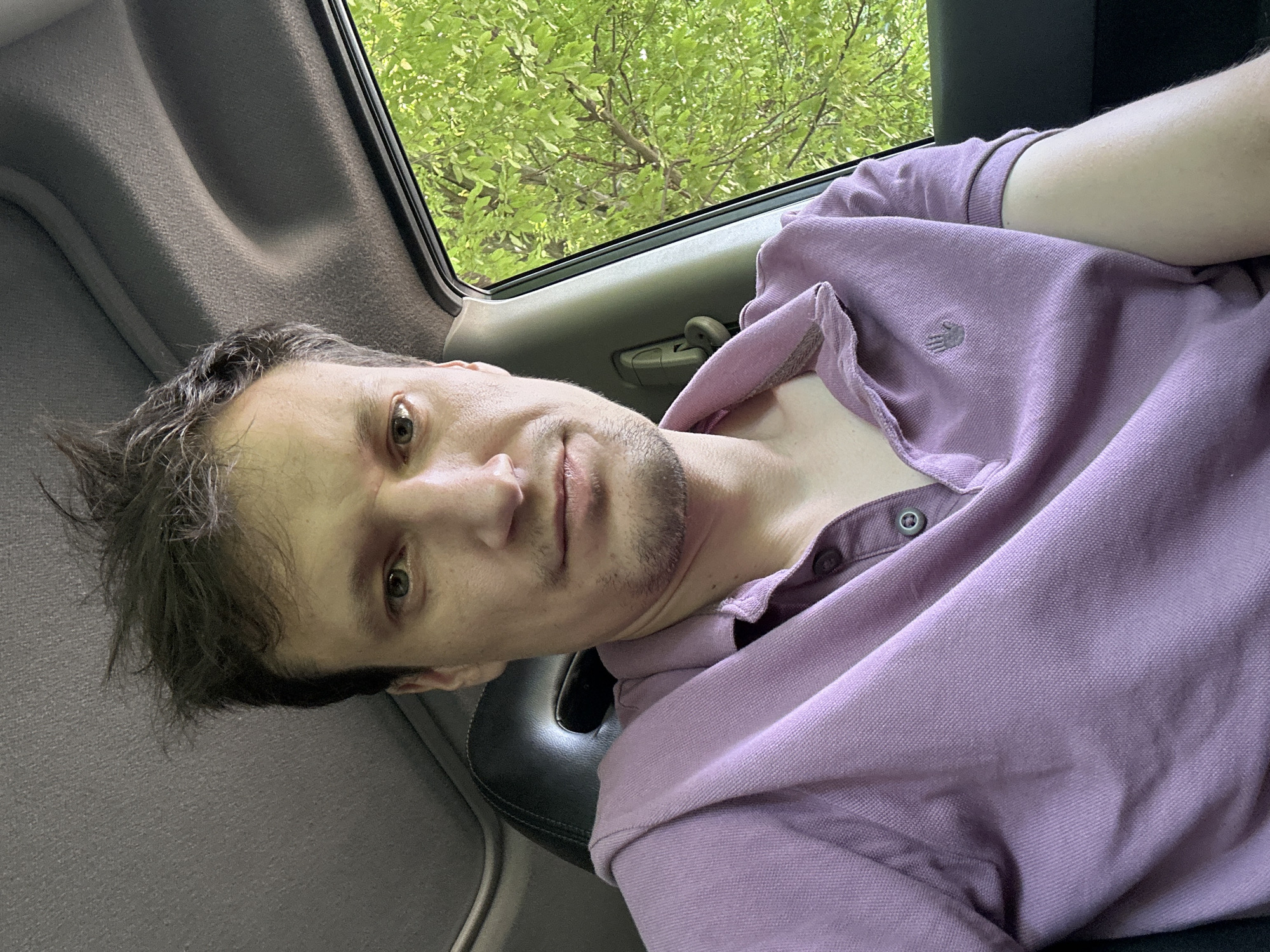 A medium shot selfie shows a man sitting in the driver's seat of a car, looking directly at the camera. He has dark, messy hair and is wearing a light purple polo shirt. Through the open car window to his right, lush green trees are visible. The interior of the car is dimly lit, with the textured fabric of the headliner and the black leather of the car seat visible. The background beyond the car is an outdoor scene with dense foliage. The man's expression is neutral, and he appears to be in a relaxed posture. The image does not provide any contextual information about the city of Shatava, Ukraine.