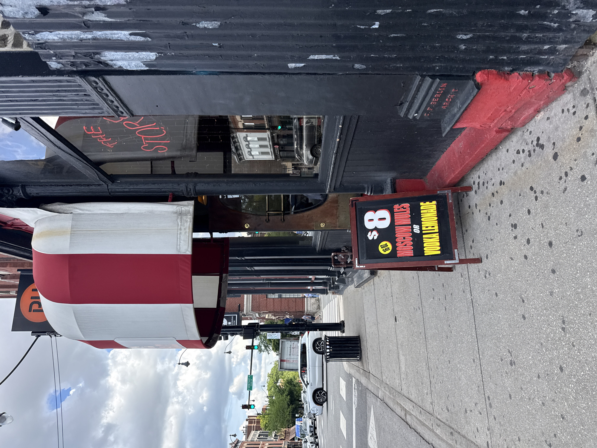 This is a photograph taken from a low angle, looking up at the exterior of a building with a red and white striped awning. The building appears to be a storefront with large windows. Red lettering on the window reads "THE STOP". A black sign with red and yellow lettering is propped up on the sidewalk in front of the building. The sign advertises "$8 MOSCOW MULES OR VODKA LEMONADE" and also lists a price of "$16.02". The sidewalk is made of concrete with visible cracks and dark spots. In the background, across the street, is a view of other buildings, including brick structures, and a street with traffic and pedestrians. The sky is cloudy, suggesting overcast weather. The overall scene is urban, likely a commercial street in Chicago, given the location context. The photograph is oriented vertically, with the top of the building appearing at the bottom of the frame.