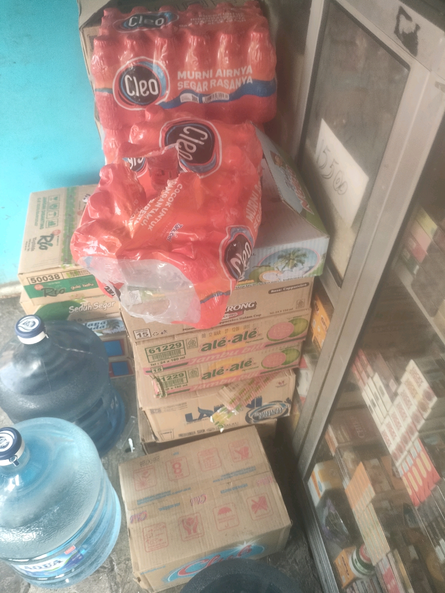 The image depicts a retail display inside a small store, likely a warung or mini-mart, located in the city of Surabaya, Indonesia. The scene is cluttered with various products, primarily beverages and packaged goods. In the foreground, two large blue water dispenser jugs are visible, one of which has a white and blue cap. Behind the jugs, stacks of cardboard boxes are arranged, containing what appear to be bottled drinks and other packaged food items. Some of the boxes display branding such as "Cleo," "Rio," and "ale-ale." In the background, a glass-fronted display cabinet contains more merchandise, suggesting a variety of consumer goods are available for purchase. The lighting is somewhat dim, and the overall impression is that of a busy, everyday retail environment. The blue wall in the background and the floor tiling suggest the location is likely an indoor space. The specific location of Surabaya is inferred from the context of the provided information, but no specific street or landmark is identifiable within the image itself.