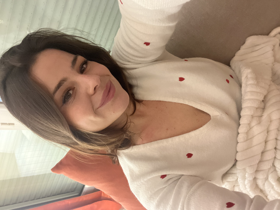 A woman with shoulder-length brunette hair smiles slightly towards the camera. She is wearing a white sweater with small red hearts scattered across it. She is resting on an orange pillow. To her right is a white, textured blanket. The angle of the photo suggests she is reclined. A window with orange trim is visible in the bottom left corner.