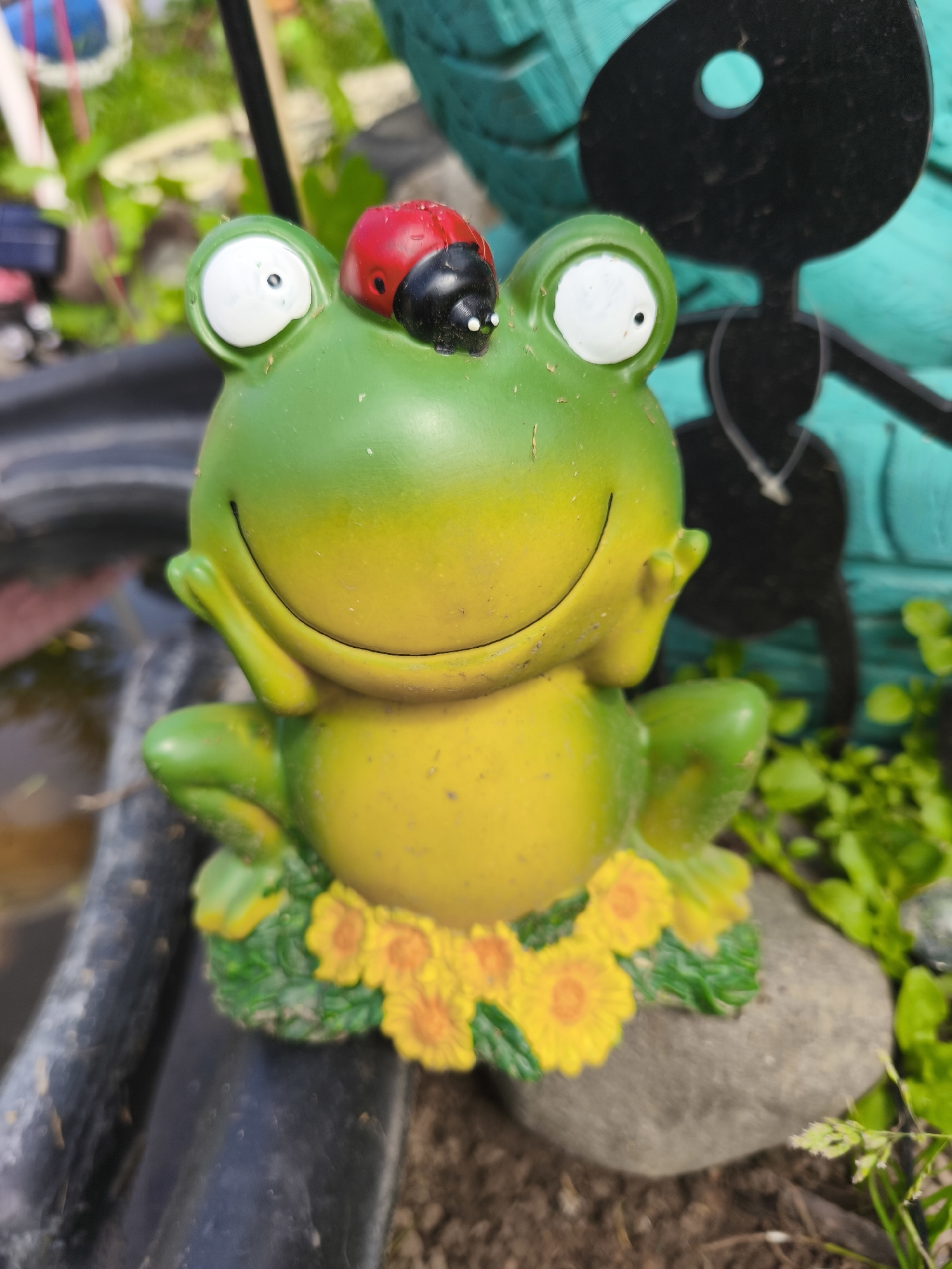 A close-up, eye-level shot focuses on a cheerful, bright green and yellow ceramic frog figurine with large, wide-set eyes. A red and black ladybug is perched on the frog's head. The frog is adorned with a ring of small, yellow, sunflower-like decorations around its base. It sits on a bed of green foliage and a grey rock, with dark soil visible in the foreground. In the background, a blurred, teal-colored object with a black silhouette of an ant-like figure is visible. The scene appears to be outdoors, possibly in a garden or park setting, with natural light illuminating the figurine. The image does not contain any elements that would suggest a specific location in Puchezh, Russia, or any other city.