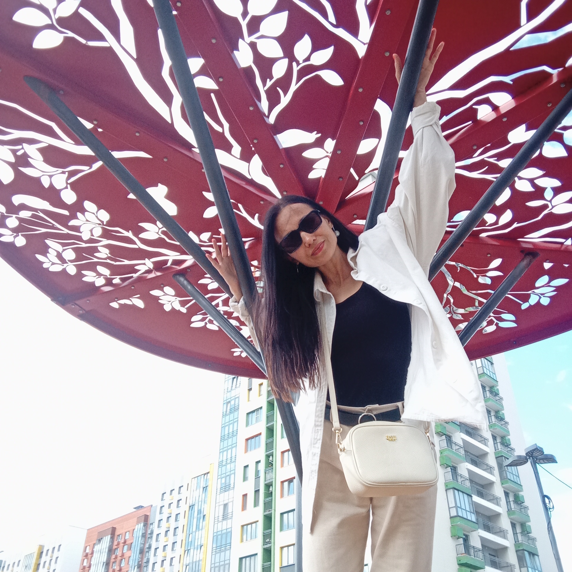 A low-angle, medium shot captures a woman standing beneath a large, red canopy decorated with a white tree and leaf motif. The woman, wearing sunglasses, a white unbuttoned shirt over a black top, and beige pants, holds one hand up, leaning against a dark gray support pole of the canopy. A small, cream-colored crossbody bag hangs from her shoulder. In the background, modern apartment buildings of varying colors, including orange, white, yellow, and green, rise into a hazy sky. A street lamp is visible on the right. The scene appears to be in an urban outdoor setting in Izhevsk, Russia.