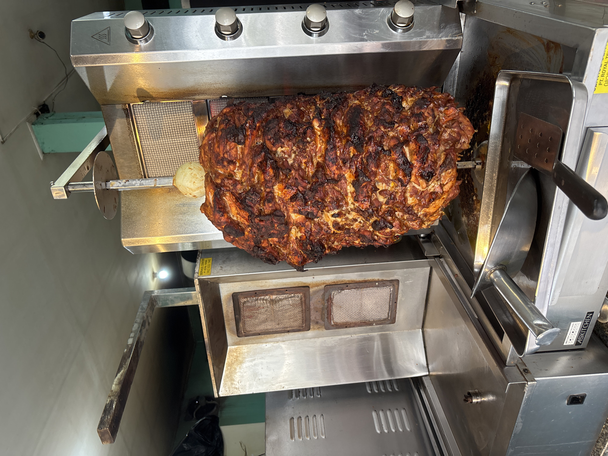 A vertical rotisserie grill stands in a commercial kitchen, its stainless-steel exterior gleaming. A large cone of roasted meat, dark and caramelized, rotates on a spit. A metal spatula rests in a tray alongside the grill, presumably used to shave the meat. The controls of the grill, including knobs, are visible at the top. A small onion sits on the spit, next to the meat. The scene is likely in a food establishment in Kuta Selatan, Indonesia, as indicated by the "SERVICE CENTER PT. ROYAL TEKNIK" label and the "ASKITCHEN" sticker.