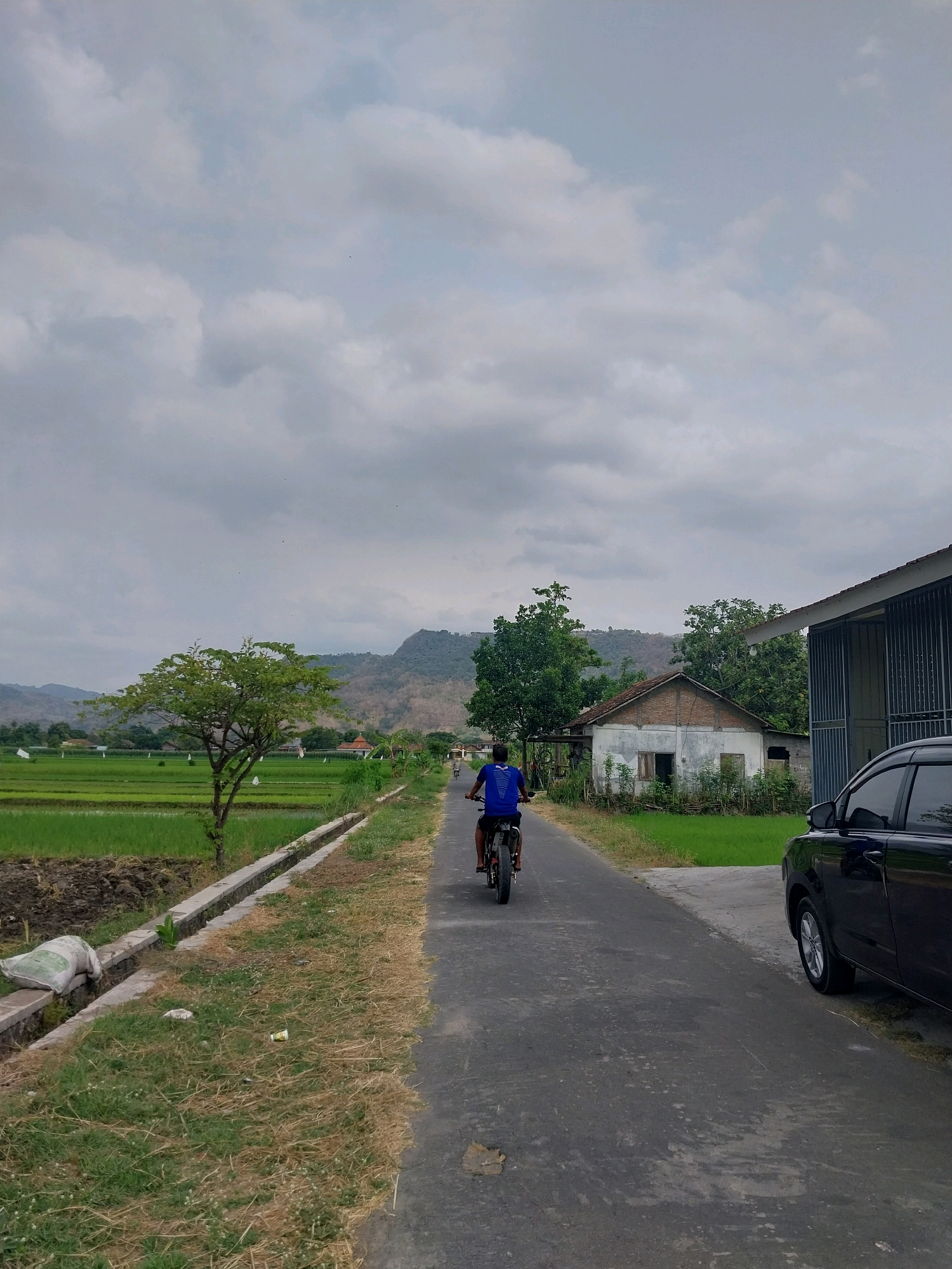 In Cucukan, Indonesia, a person is riding a motorbike down a paved road. The road is flanked by a lush green rice field on the left and a small, weathered house with a red-tiled roof on the right. Beyond the house, a modern building with a metal fence stands. In the distance, rolling hills are visible under a cloudy sky. A black car is parked on the right side of the road. A solitary tree with green foliage stands in the middle ground of the rice field.