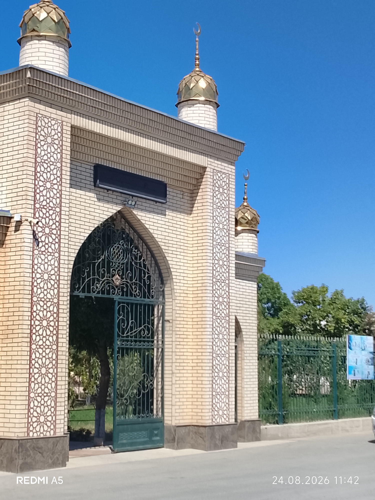 The image shows an exterior view of a mosque in Pakhtakor, Uzbekistan. The mosque features light-colored brickwork, decorative patterned panels on its facade, and ornate golden domes topped with crescent moons. An arched gateway with intricate green metalwork is partially open, revealing a glimpse of trees and greenery within the mosque grounds. A green metal fence runs along the side of the property, with a sign visible on it. The sky is clear and blue. A timestamp in the bottom right corner reads "24.08.2026 11:42," and "REDMI A5" is visible in the bottom left corner. There are no people visible in the image.