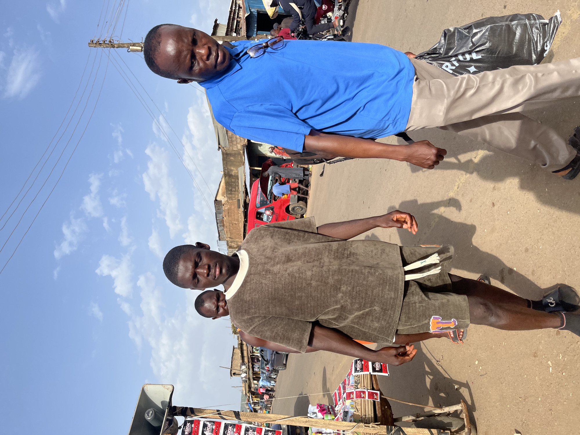 In Rafin Par, Nigeria, two men walk along a dusty, sunlit street lined with shops and market stalls. The man in the foreground on the right wears a bright blue polo shirt and light brown pants, carrying a black plastic bag in his right hand. To his left, another man, closer to the camera, wears a brown, corduroy t-shirt and patterned shorts. Behind him, a third man in the background wears an orange shirt. The street is populated with other individuals and vehicles, including a red van with its hood open. To the left of the frame, a market stall displays various products on shelves and a large speaker system is mounted above. The sky is blue with scattered white clouds.