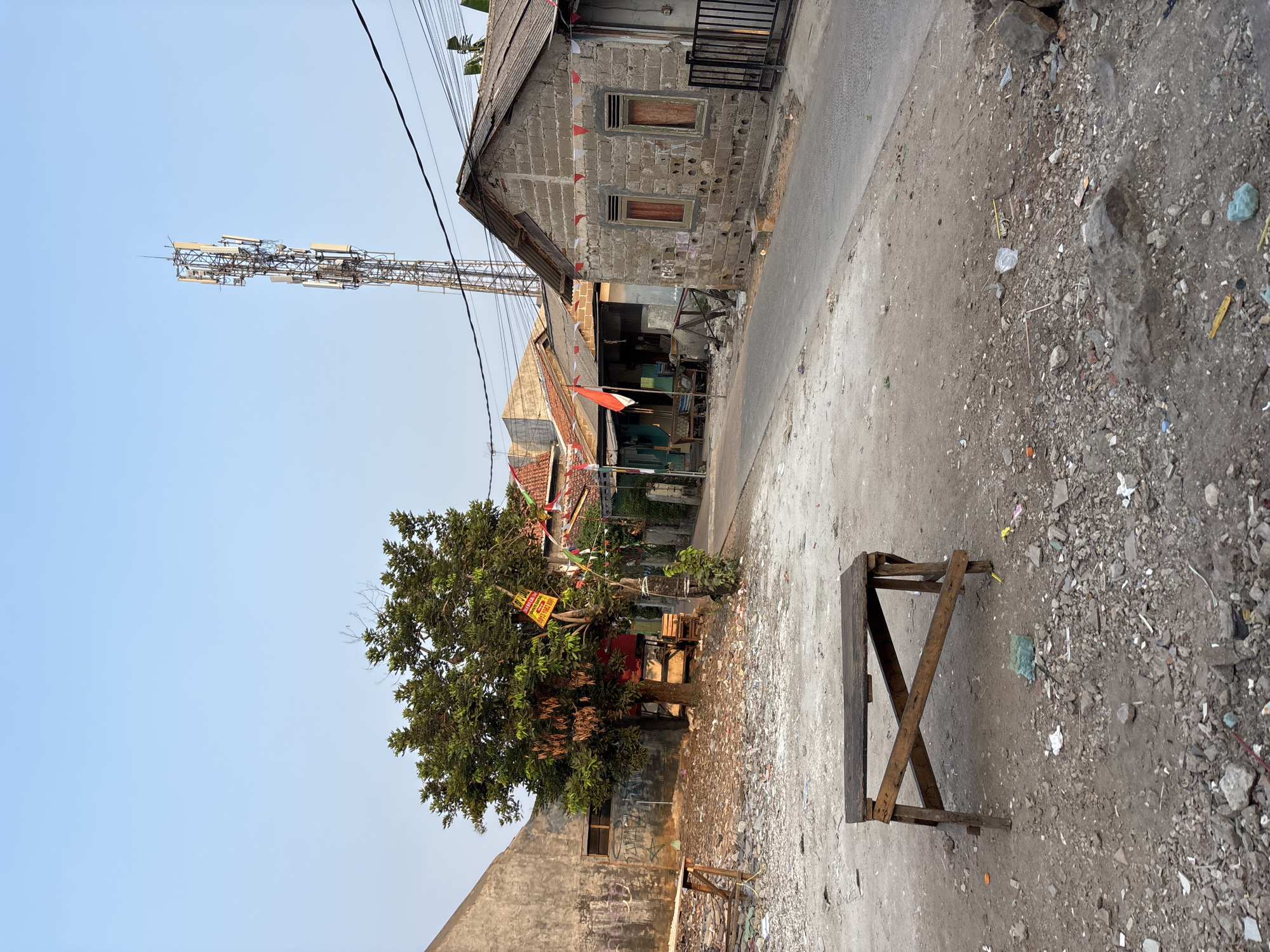 The image depicts a street scene in Bekasi, Indonesia, characterized by a clear sky and a somewhat dilapidated environment. In the foreground, a rustic wooden table with a simple X-shaped support structure sits on a ground covered with gravel, dirt, and scattered debris, including plastic waste and small rocks.
To the left, a building with a textured concrete wall, exhibiting some graffiti, stands partially visible. Adjacent to it is a leafy green tree adorned with Indonesian flags and pennants, suggesting a celebration or national holiday. Behind the tree, more structures and a red-colored element are visible, possibly a stall or small building.
The middle ground reveals a paved road leading into a residential area. On the right side of the road, a modest house constructed from concrete blocks is prominent. It has a corrugated metal roof, two windows with red curtains, and a metal fence in front. Several other houses and buildings are visible further down the street, some also decorated with flags.
In the background, a tall telecommunications tower with multiple antennas dominates the skyline, an indication of modern infrastructure amidst the more traditional or less developed surroundings. Power lines crisscross the sky above the buildings. The overall impression is one of a working-class neighborhood in Bekasi, with elements of both everyday life and national pride evident.
