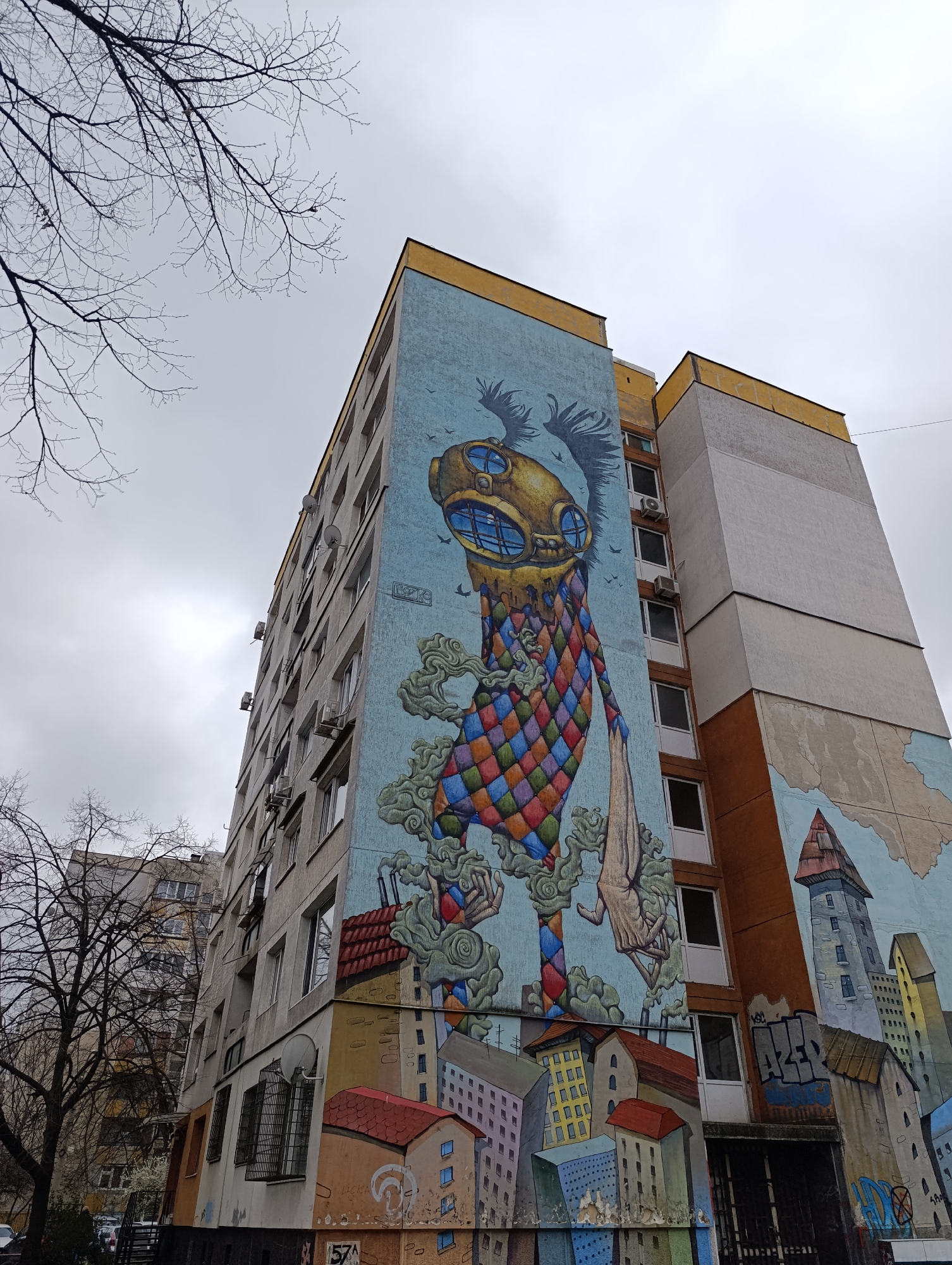 The image shows a tall, grey apartment building in Sofia, Bulgaria. A large, colorful mural covers one side of the building. The mural depicts a cartoonish figure wearing a diving helmet and a harlequin suit. The figure is surrounded by smoke and stands above a cityscape painted on the lower part of the building. The building has numerous windows and air conditioning units. Tree branches are visible in the foreground against a cloudy sky. Another building is partially visible to the left of the apartment building. There are some small street building murals as well.