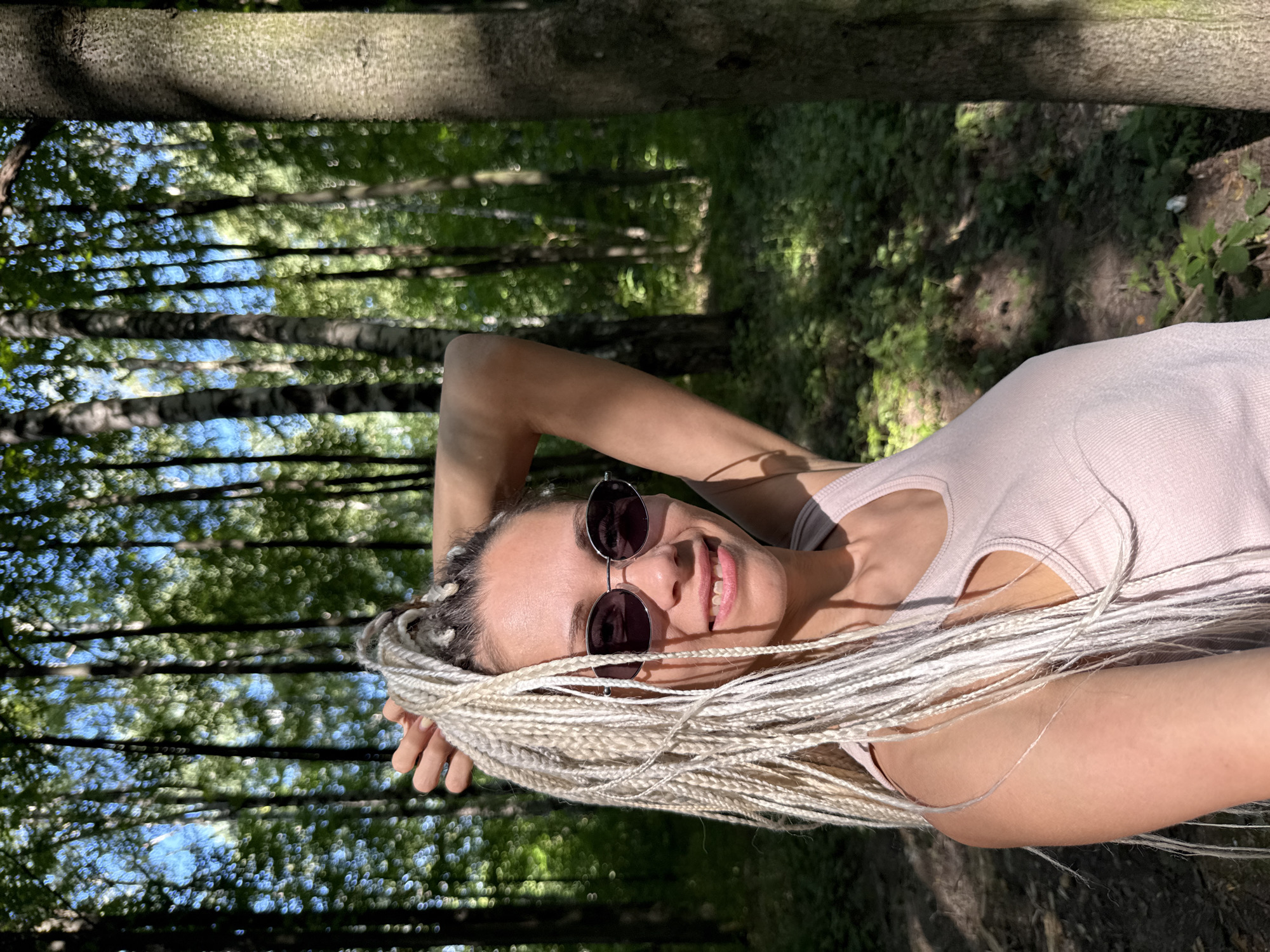 A woman with long, blonde braided hair and sunglasses smiles at the camera while standing in a forest with tall trees. She is wearing a light pink tank top. Her left arm is raised, with her hand in her hair. Sunlight filters through the leaves, creating dappled shadows on the forest floor. The location is identified as Rusanovo, Russia.