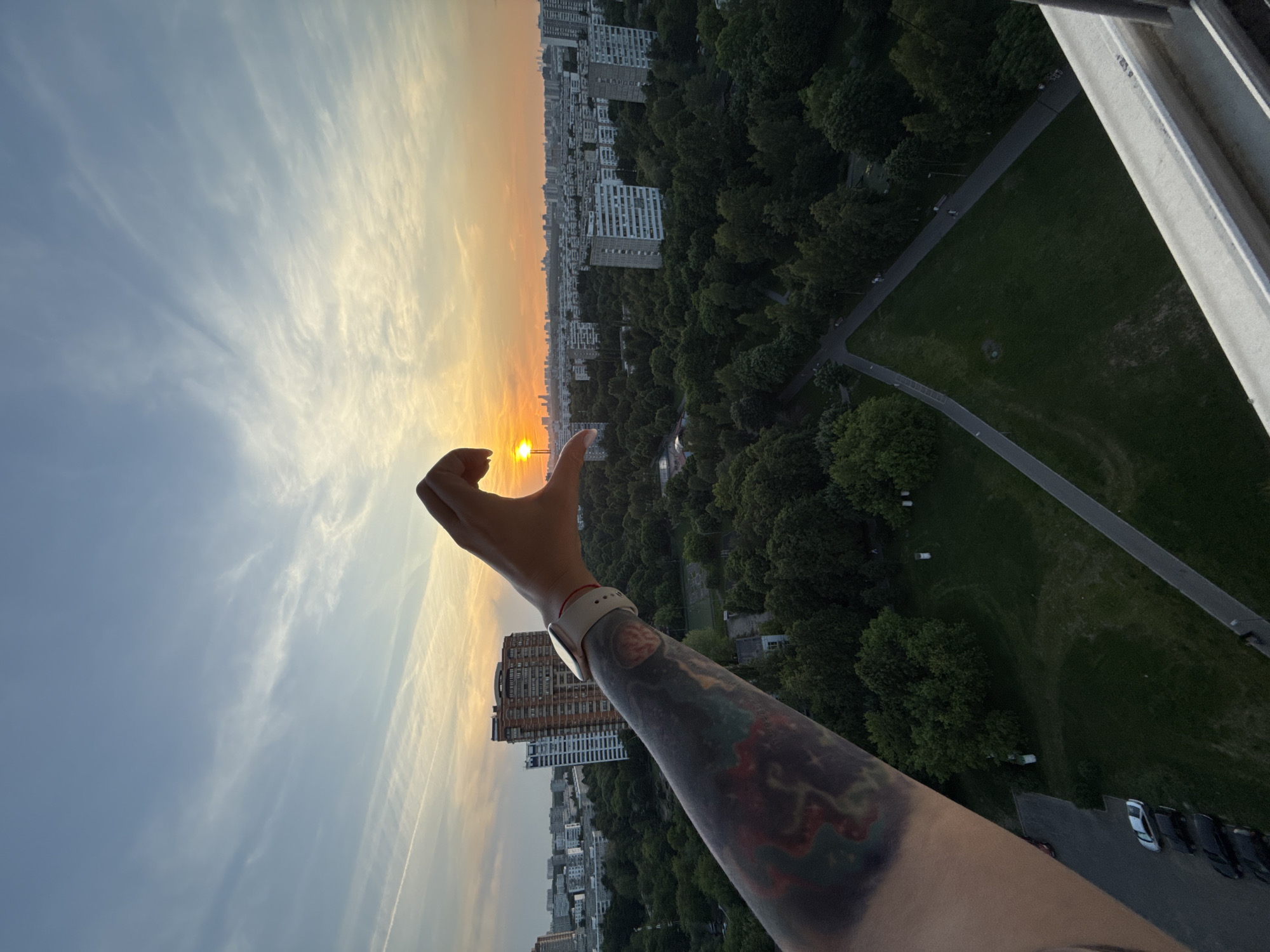 The image displays a high-angle view of a residential district in Moscow, Russia, specifically identified as the Khoroshevo-Mnevniki District, captured during sunset. In the immediate foreground, a person's right arm, heavily tattooed with abstract, multi-colored designs, extends from the lower-left. A light-colored smartwatch with a visible screen and a thin red string are worn on the wrist. The hand is positioned to form a half-heart shape, framing the bright yellow-orange setting sun between the thumb and index finger.
The urban environment below is densely populated with multi-story residential buildings. A prominent, large, multi-section reddish-brown apartment complex, featuring horizontal white accents, is visible in the mid-left background, adjacent to a taller, slender white building characterized by vertical lines. Numerous typical Soviet-era panel buildings with repetitive window patterns are scattered across the middle ground, extending towards a distant, flatter city skyline on the horizon.
Extensive green spaces, comprising large parks and tree-lined areas, are interspersed throughout the cityscape. In the lower-mid ground, a large park features paved pedestrian walkways, one leading diagonally from the bottom-left and another from the bottom-right. Several white cylindrical or cubic objects are visible within the park. A small group of parked cars is partially visible on an asphalt lot in the lower-left. The sky exhibits warm orange and yellow hues at the horizon, transitioning to light blue in the upper portion, with wispy white cirrus clouds and a single aircraft contrail. A white window sill or balcony railing is visible along the bottom edge of the frame, indicating the elevated vantage point of the photographer.