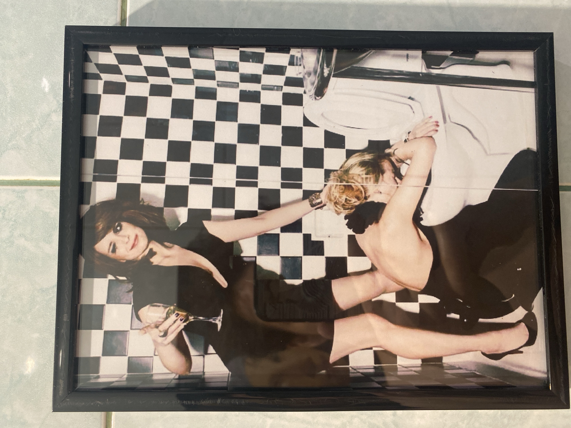 The image shows a framed photograph. The photograph depicts two women in a bathroom with a black and white checkered floor. One woman is lying down, holding a champagne glass. The other woman is leaning over a toilet. The location is not discernible from the image.