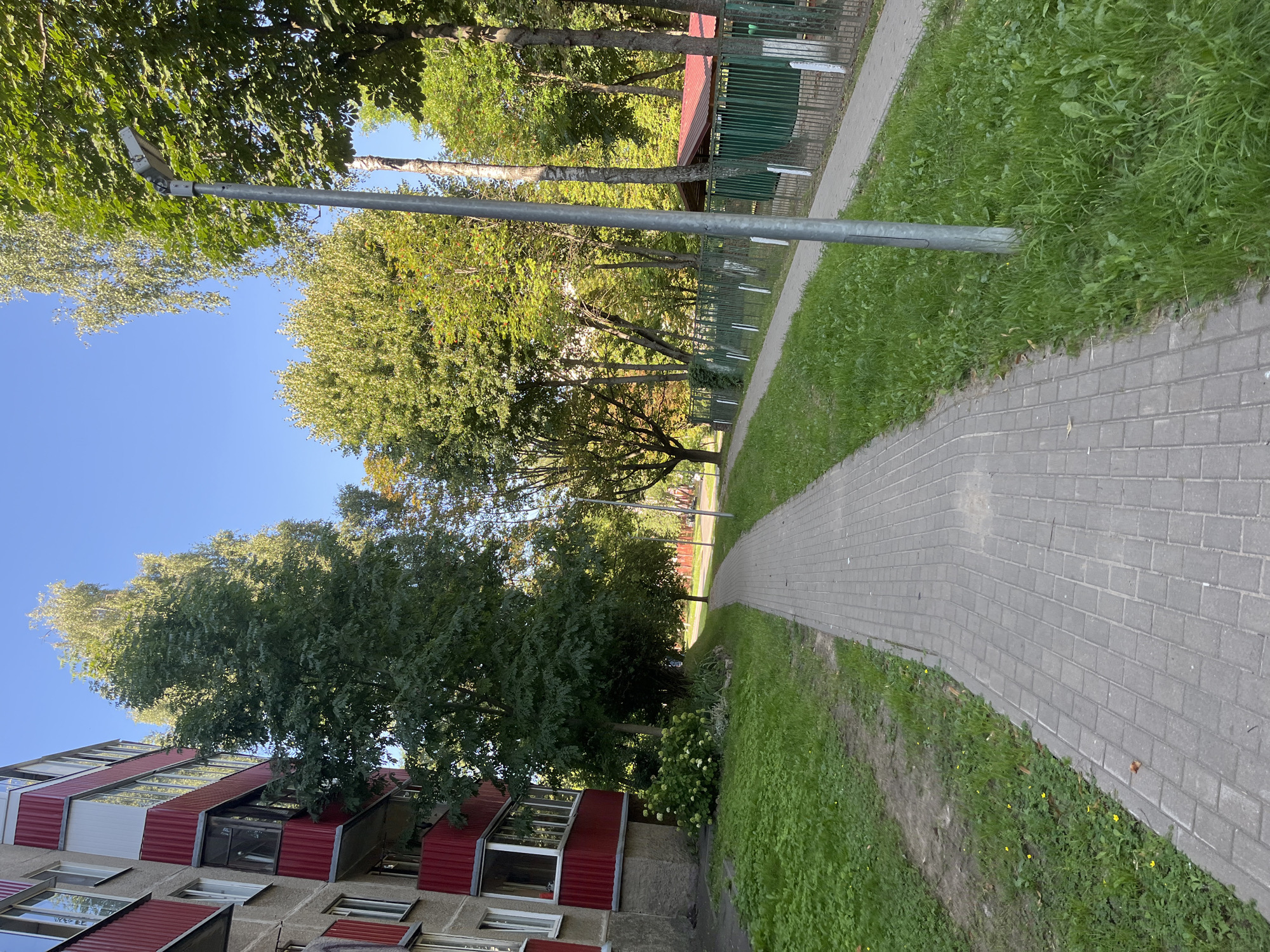 The image shows a paved walkway in a residential area, likely in Minsk, Belarus, given the architectural style of the building and the presence of deciduous trees commonly found in the region. The walkway is made of grey interlocking pavers and runs alongside a grassy area and a multi-story apartment building with red balconies. The building's facade is primarily concrete, with red and white elements on the balconies. To the right of the walkway, there is a chain-link fence, behind which is a low-rise structure with a red roof and green walls, possibly a utility shed or small building. A lamppost stands beside the walkway. The environment is characterized by lush green grass, numerous trees with dense foliage, and a clear blue sky. There are no people or animals visible in the frame. The scene is bathed in daylight, suggesting it was taken during the day. The overall impression is one of a quiet, residential street with urban greenery.