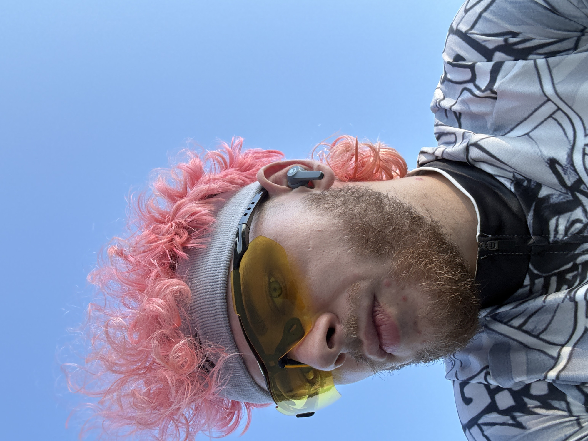 A low-angle close-up shot shows a person with light skin, a reddish-brown beard, and pink curly hair. They are wearing a light gray beanie and yellow-tinted sunglasses. A gray earbud is visible in their left ear. The person is wearing a patterned white and black garment. The background is a clear blue sky. The location in Đà Nẵng, Vietnam is not discernible from the image.