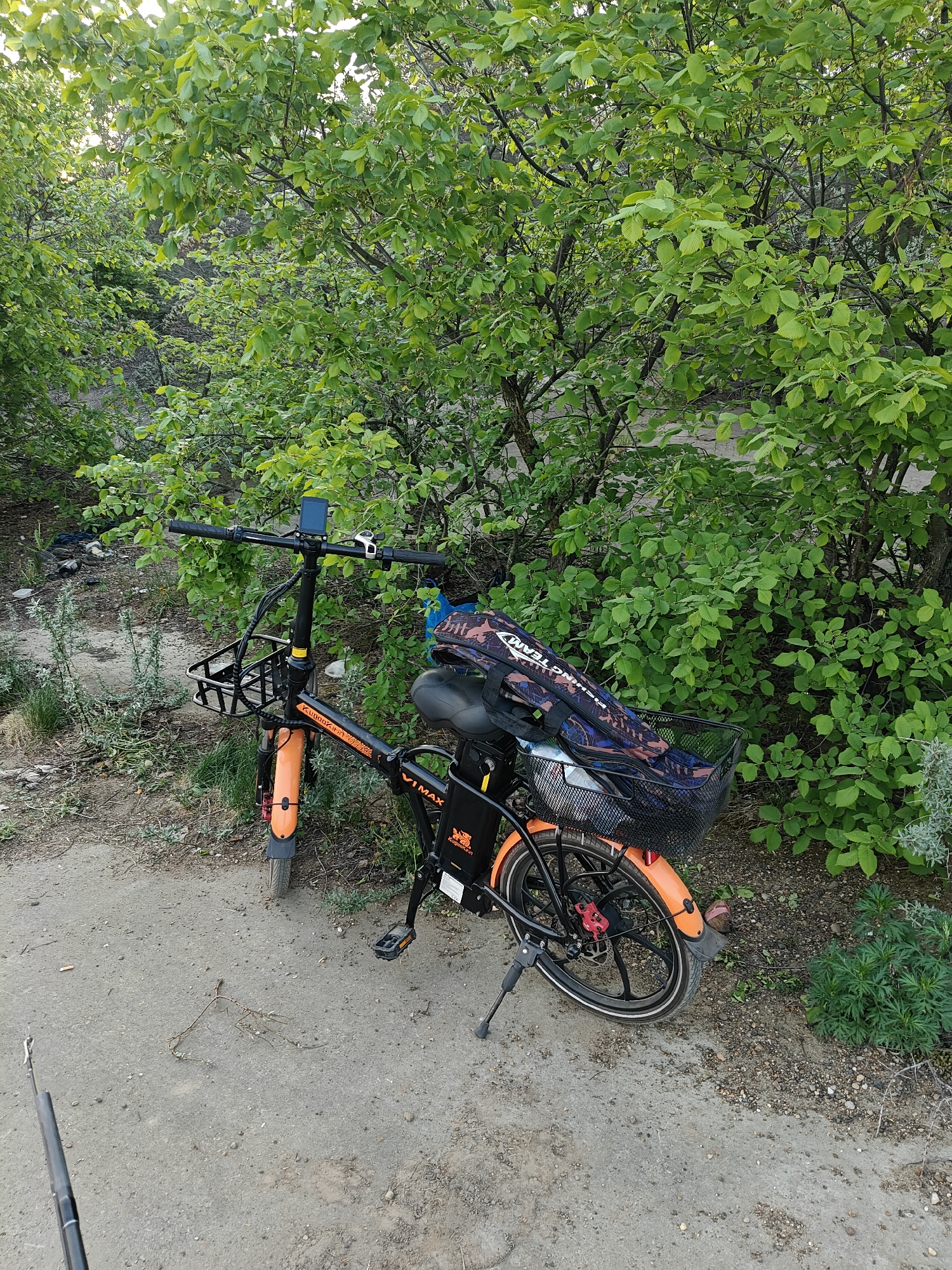 The image shows a black and orange folding electric bicycle parked on a dirt and gravel path in an outdoor environment. A black basket is attached to the front of the bicycle, and a fishing rod case rests in a rear basket. The bicycle is equipped with a kickstand, supporting it upright. Green leafy bushes fill the background behind the bicycle. A portion of a fishing rod is visible at the lower left corner of the image. The location is Chaikovsky, Russia.