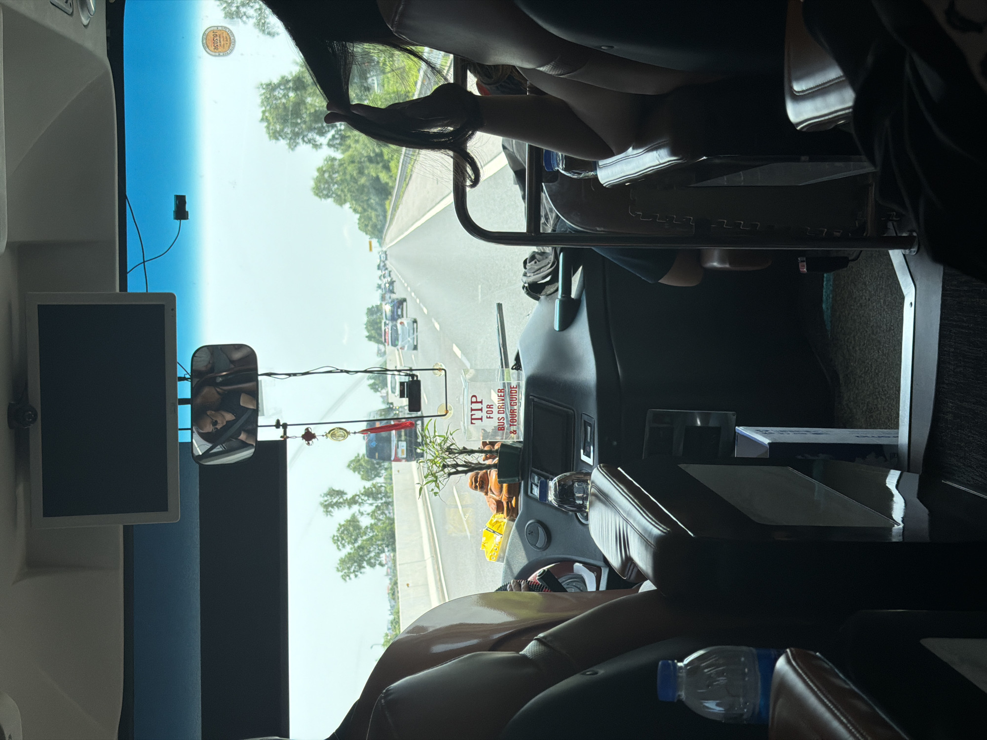The image captures the interior of a bus or coach, looking forward through the windshield onto a road. The perspective is from a passenger seat, with a person's arm and long dark hair visible in the foreground, seemingly holding a phone. A plastic water bottle rests next to the seat.
The dashboard features various controls, a built-in screen, and a rearview mirror reflecting a person's face. Decorative items are placed on the dashboard, including a small potted plant, a golden Buddha statue, and red and gold hanging ornaments. A white sign with prominent red text reads "TIP FOR BUS DRIVER & TOUR GUIDE."
To the left, a monitor is mounted, and a dashcam or small camera is attached to the upper part of the windshield. A circular sticker or permit is also affixed to the windshield, showing numbers that are partially legible (e.g., "105030").
Beyond the windshield, a multi-lane highway or main road stretches into the distance, with several cars and trucks visible, indicating traffic. The weather appears clear and sunny, with a bright blue sky and green trees lining the road. The scene suggests daytime travel on a busy road in Hải Phòng, Vietnam.