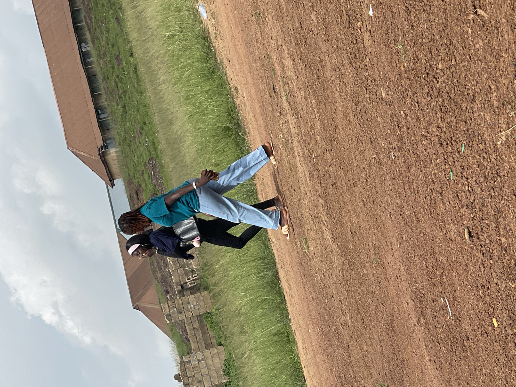 Two young women are walking on a dirt road. The woman on the left is wearing a dark blue headscarf, a dark blue top, and black pants. She is carrying a black purse. The woman on the right is wearing a teal t-shirt and light blue jeans. She is wearing sandals. They are walking past a grassy area with some dilapidated brick structures on the left and a building with a rusty, corrugated metal roof on a hill in the background. The sky is overcast with white clouds. The scene is likely in a rural or undeveloped area outside of a city in Nigeria, given the visual cues of the environment.