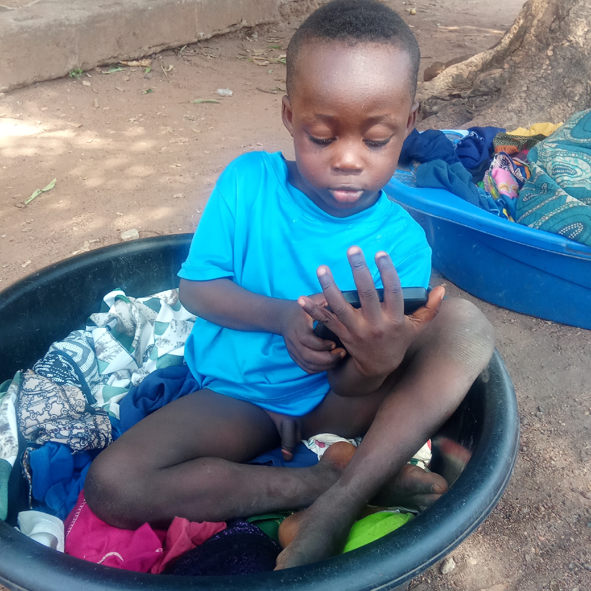 A young Black child sits cross-legged in a black plastic basin filled with colorful clothing. He is wearing a bright blue T-shirt and holds a dark-colored mobile phone in his hands, appearing to be focused on its screen. The basin is placed on a reddish-brown, dirt surface. To the right, a blue plastic container holding additional textiles is visible. The ground suggests an outdoor setting, with a tree trunk partially seen in the background. The location is identified as Akwanga, Nigeria.