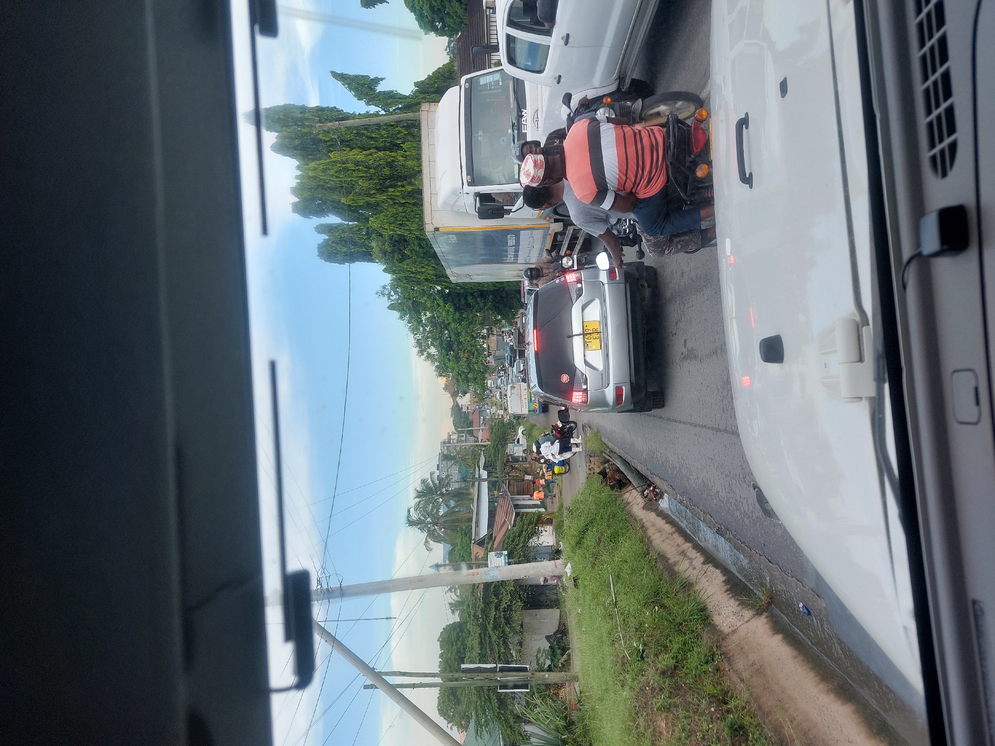 The image shows a street scene in Dar es-Salaam, Tanzania, likely a traffic jam. The view is from inside a vehicle, looking out onto a busy road. Several vehicles are visible, including a white truck, a silver car with a license plate ending in "EIR," and numerous motorcycles weaving through the traffic. Pedestrians are also present along the roadside. The environment consists of a mix of urban and natural elements, including buildings, trees, and a grassy roadside area. Overhead power lines are also noticeable. The weather appears clear and sunny.