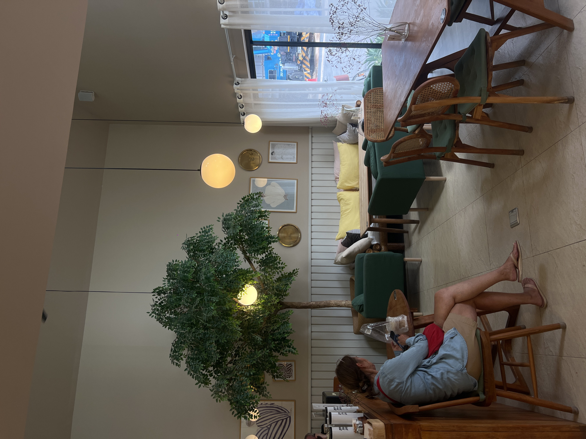 Here is a description of the image:
**Scene:** The image depicts the interior of what appears to be a cafe or restaurant. The style of the establishment is modern and airy, with a focus on natural elements.
**Location:** Unknown city, Unknown.
**People:** One visible person is seated in a chair, holding a smartphone. The person is wearing shorts, a red shirt, and a denim jacket. Their legs are propped up on another chair.
**Objects:** A large faux tree occupies a significant portion of the space. The furniture includes green upholstered chairs, a wooden table, and a sofa with yellow pillows. There are also decorative items on the wall, such as framed art and a mirror. Coffee cups are also visible in the corner.
**Environment:** The cafe has large windows that allow natural light to enter. A construction site with machinery is visible through the window. The lighting is soft and ambient, with globe-shaped hanging lights.