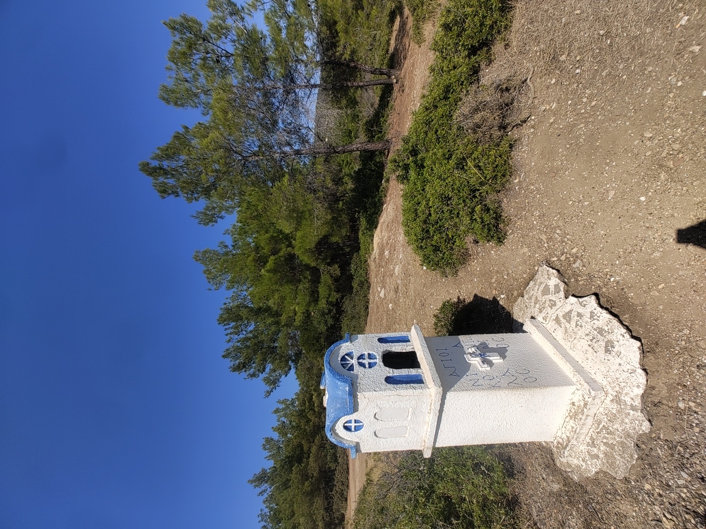 A small, white, church-shaped structure with a blue roof and blue trim stands on a rocky base in a dry, outdoor environment. The structure has arched windows and a cross on its front. Greek text is visible on the front of the structure. The background consists of a dirt path, green shrubs, and numerous pine trees under a clear blue sky. The location is identified as Limenaria, Greece.