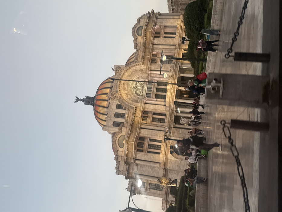 An ornate building stands tall, its dome striped in hues of orange and gold. The structure, possibly a palace or museum, is a blend of neoclassical and Art Deco styles. People gather in the foreground. A chain fence is blurred, adding depth. The sky is overcast.