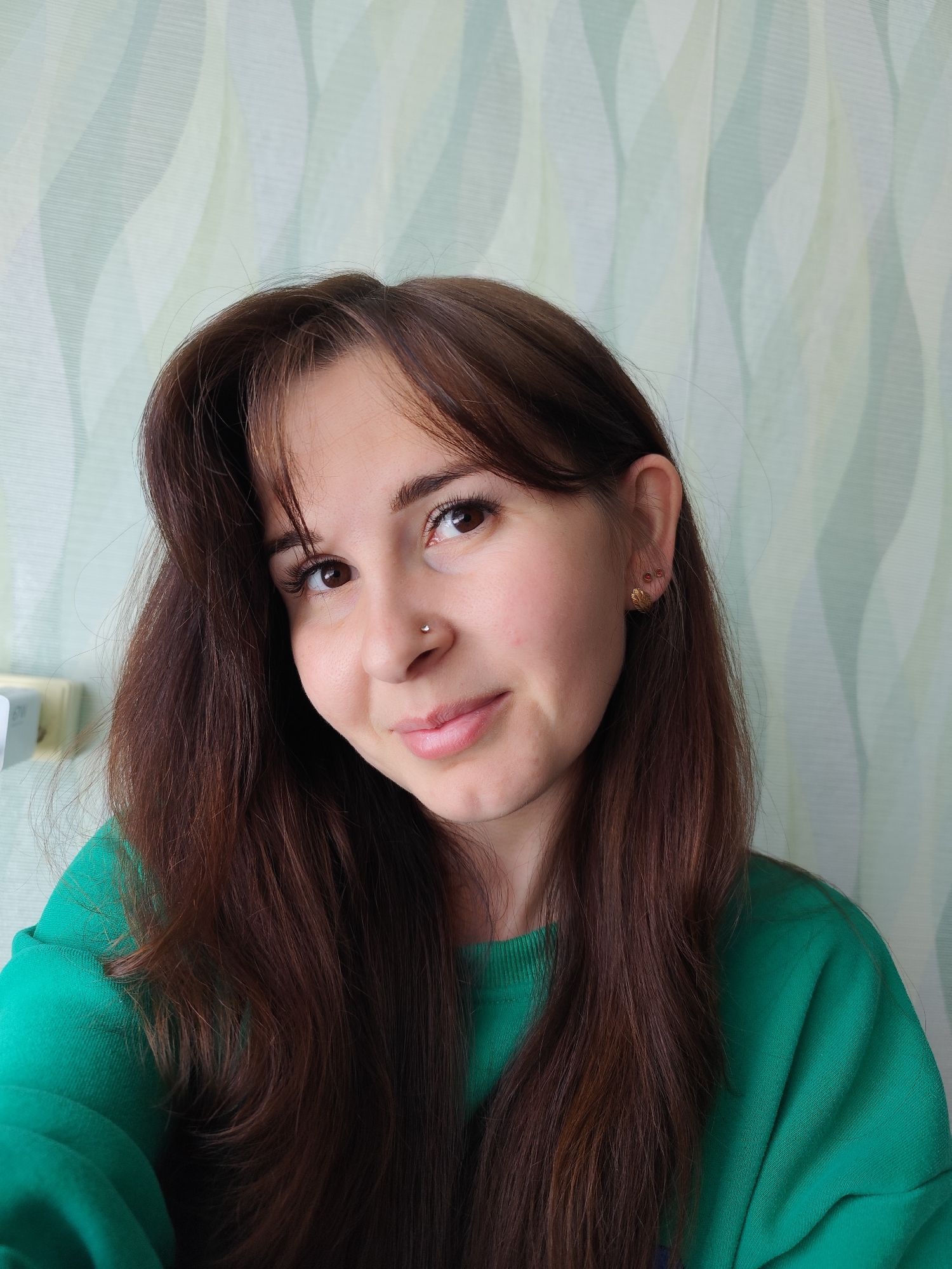 A young woman is featured in a close-up, chest-up shot, looking directly at the camera with a soft, inviting smile. She has long, dark brown hair styled with bangs, which falls over her shoulders and down her front. Her eyes are brown, and her eyelashes appear prominent. A small, silver stud piercing is visible on her right nostril. She is wearing a vibrant emerald green crew-neck sweatshirt. In her right ear, she has two golden earrings: a decorative stud shaped like a leaf or wing, and a smaller, plain stud above it.
The background consists of wallpaper featuring a subtle, wavy vertical pattern in shades of light green and cream or white, creating a gentle, flowing aesthetic. On the far left side of the image, a partial view of a white electrical outlet or light switch is visible against the wall. The lighting suggests an indoor setting, likely during the daytime, casting an even glow on her face. No discernible text is visible within the image. The scene conveys a relaxed and casual atmosphere, captured in Фоки, Russia.
