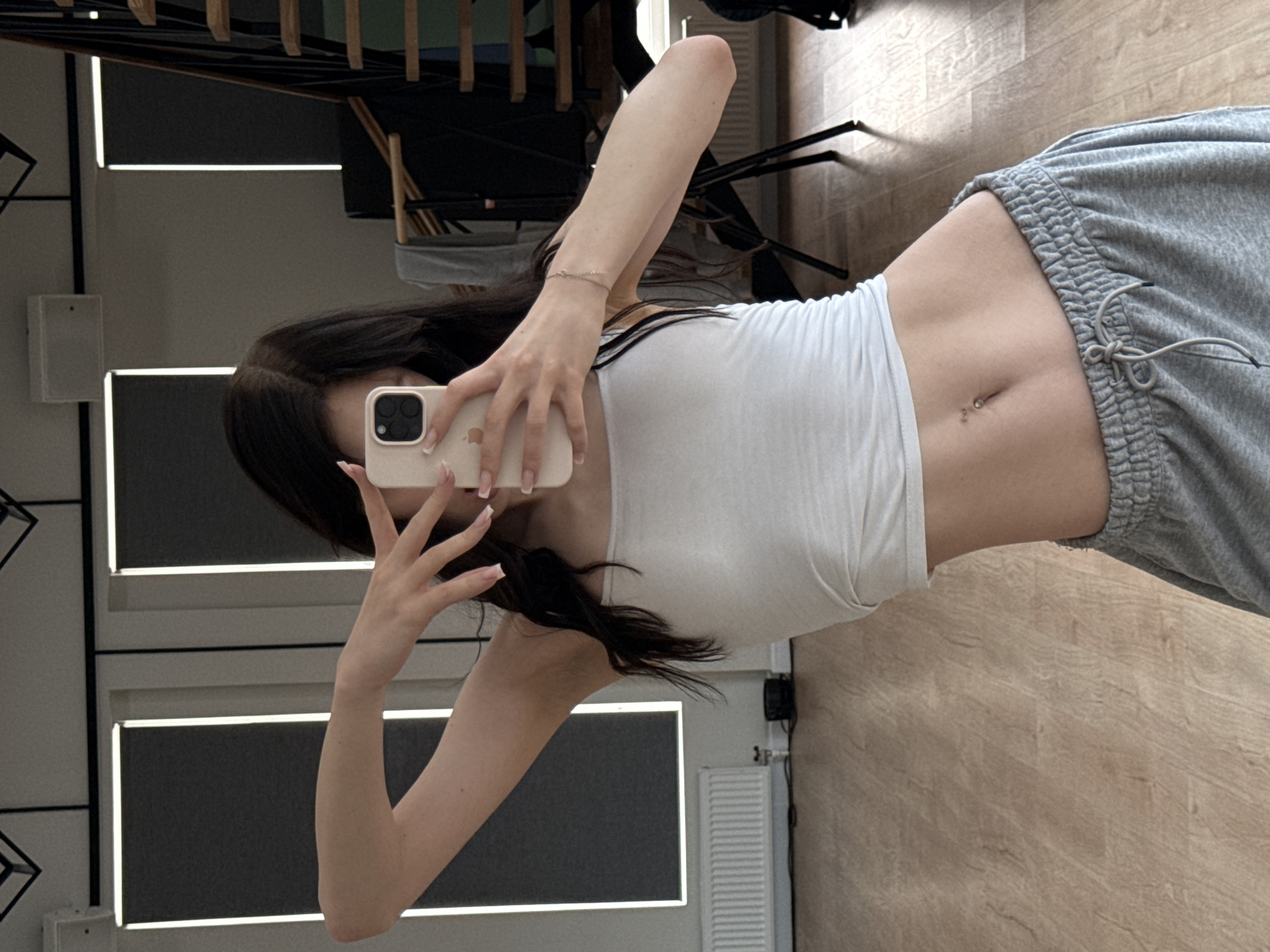 A young woman is taking a mirror selfie in a room with light wood flooring. She has long, dark hair and is wearing a white tank top and grey sweatpants. Her midriff is exposed, showing a pierced belly button. She is holding a smartphone in her right hand to take the picture. The background includes a mirror, some furniture, and a radiator. The lighting suggests it is daytime, and there are no clear indicators of the weather. The overall mood appears casual and relaxed. No visible text is present in the image.