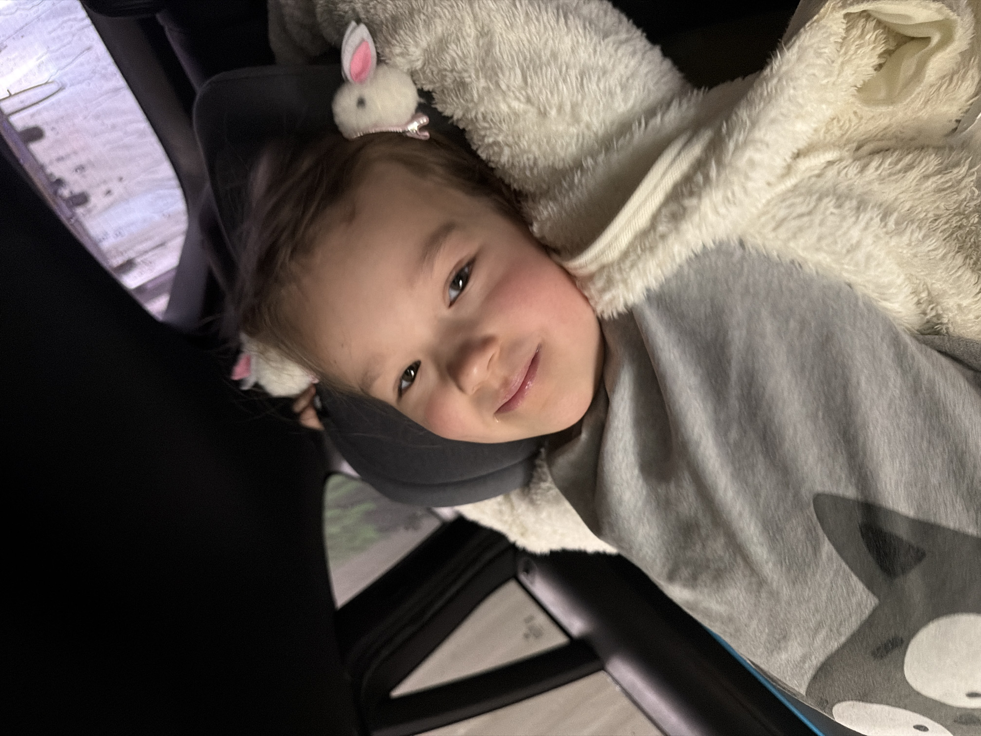 A young child with light brown hair and a rabbit-shaped hair clip is resting in a car. She is wearing a gray shirt with a wolf design and is partially covered by a fluffy, off-white blanket. The child is reclining against a dark gray neck pillow, and parts of the car interior, including the seat and window, are visible. The location within the city of Saint Petersburg, Russia, cannot be determined from the image.
