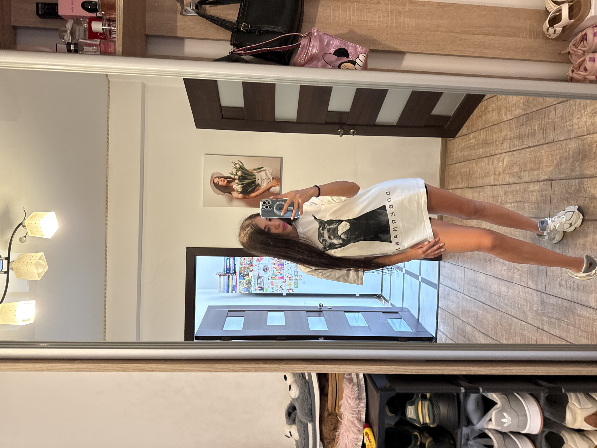 A young woman stands in front of a full-length mirror, taking a selfie. She has long, dark hair and is wearing a white t-shirt with a graphic print of a dog's head and text, and dark shorts. Her reflection shows her holding a smartphone in her right hand. The mirror is in a room with light-colored walls and wooden flooring. To the left of the mirror, a shoe rack is visible, stacked with various shoes and slippers. On the right side of the mirror, a wooden closet with shelves displays perfumes and bags, and a framed picture of a woman holding tulips hangs on the wall. A partially open door leads to another room, revealing shelves filled with various items. The overall scene suggests a domestic setting, likely a bedroom or hallway within a residential apartment in Chisinau, Moldova, given the context provided.