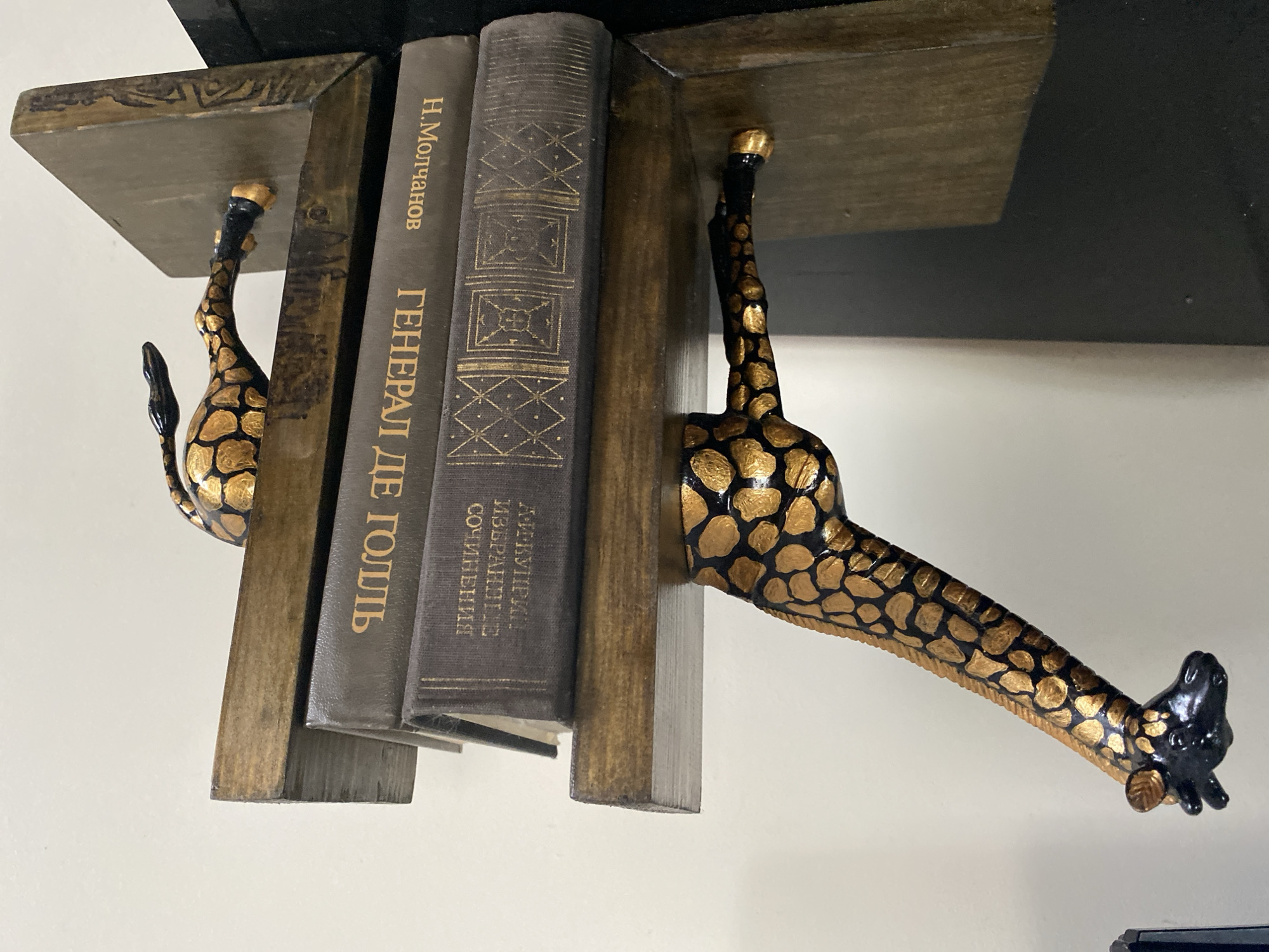 The image shows a pair of decorative bookends shaped like giraffes. One giraffe is positioned on the left, with its body curving around to hold a stack of two books. The giraffe is black with gold spots and gold hooves. The second giraffe is on the right, also black with gold spots, and appears to be holding the other side of the book stack, though only its hindquarters and tail are visible. The books are dark grey with gold lettering and decorative patterns on their spines. The bookends and books are placed on a dark surface, possibly a shelf or table, in front of a light-colored wall. The overall impression is one of a decorative display. There is no information in the image that indicates the location in the city of Vitsebsk, Belarus.