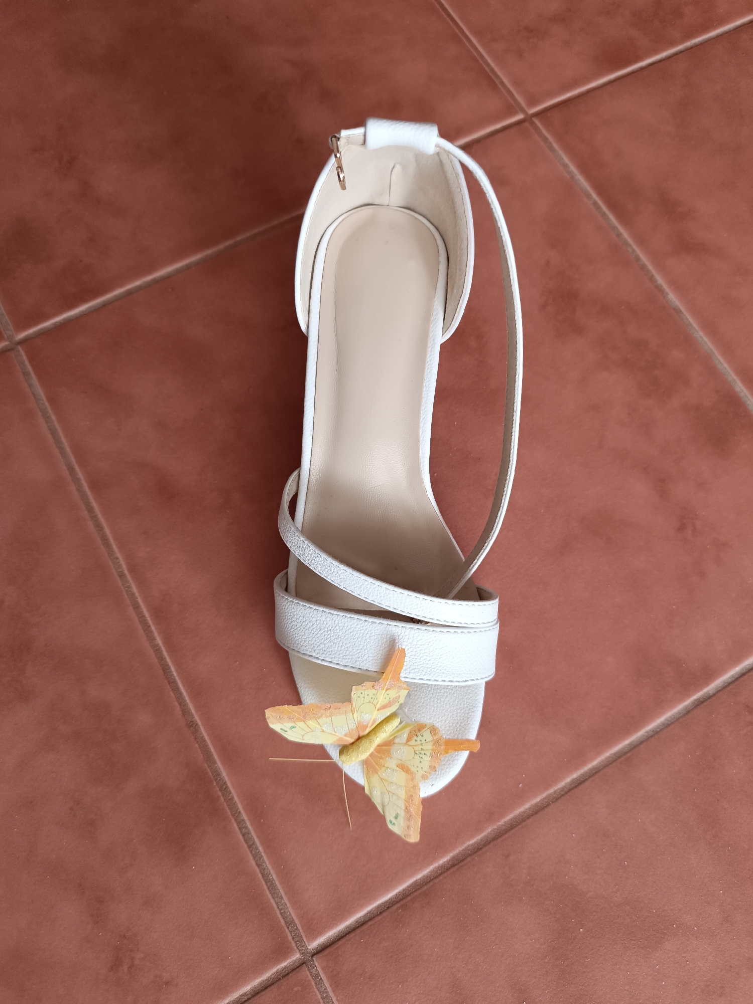 A white heeled sandal with crisscrossing straps and a butterfly embellishment is positioned on a terracotta tiled floor. The sandal is oriented vertically with the toe pointing downwards. The butterfly, in shades of yellow and orange, is placed on the front strap of the sandal. The background consists of a grid of brown square tiles, with visible grout lines. The scene is indoors, likely a home environment. There are no people present in the image. The lighting suggests a bright, possibly natural light source, casting subtle shadows. The image is a top-down view.