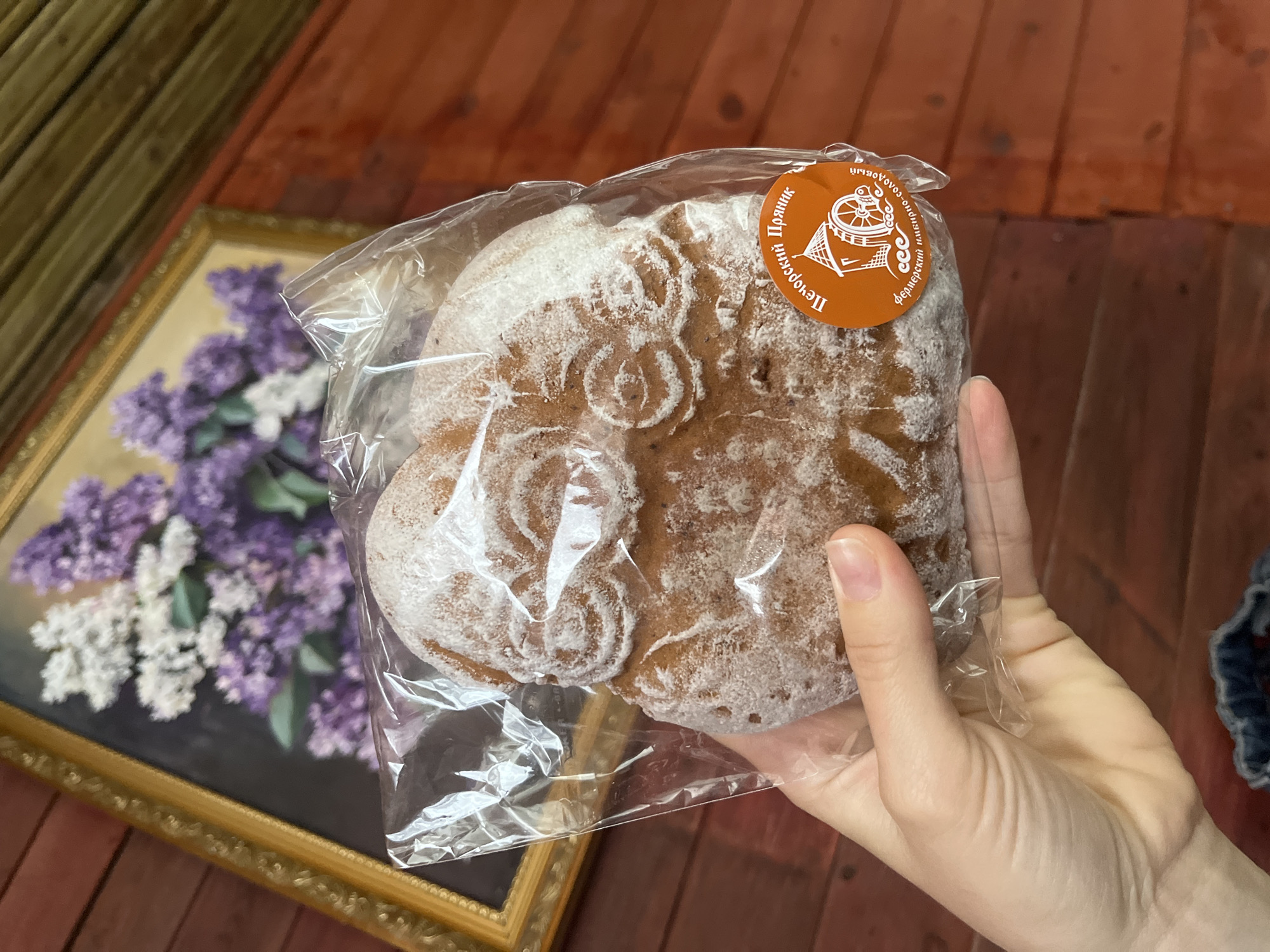 A hand holds a cellophane-wrapped, gingerbread-like pastry, likely in the city of Мшинская, Russia, based on the Russian text on the circular sticker. The pastry is covered in a white powder, possibly powdered sugar, and has decorative designs. The background includes a framed painting of lilacs and a dark wood surface. A small portion of what appears to be denim clothing is visible in the background on the right.