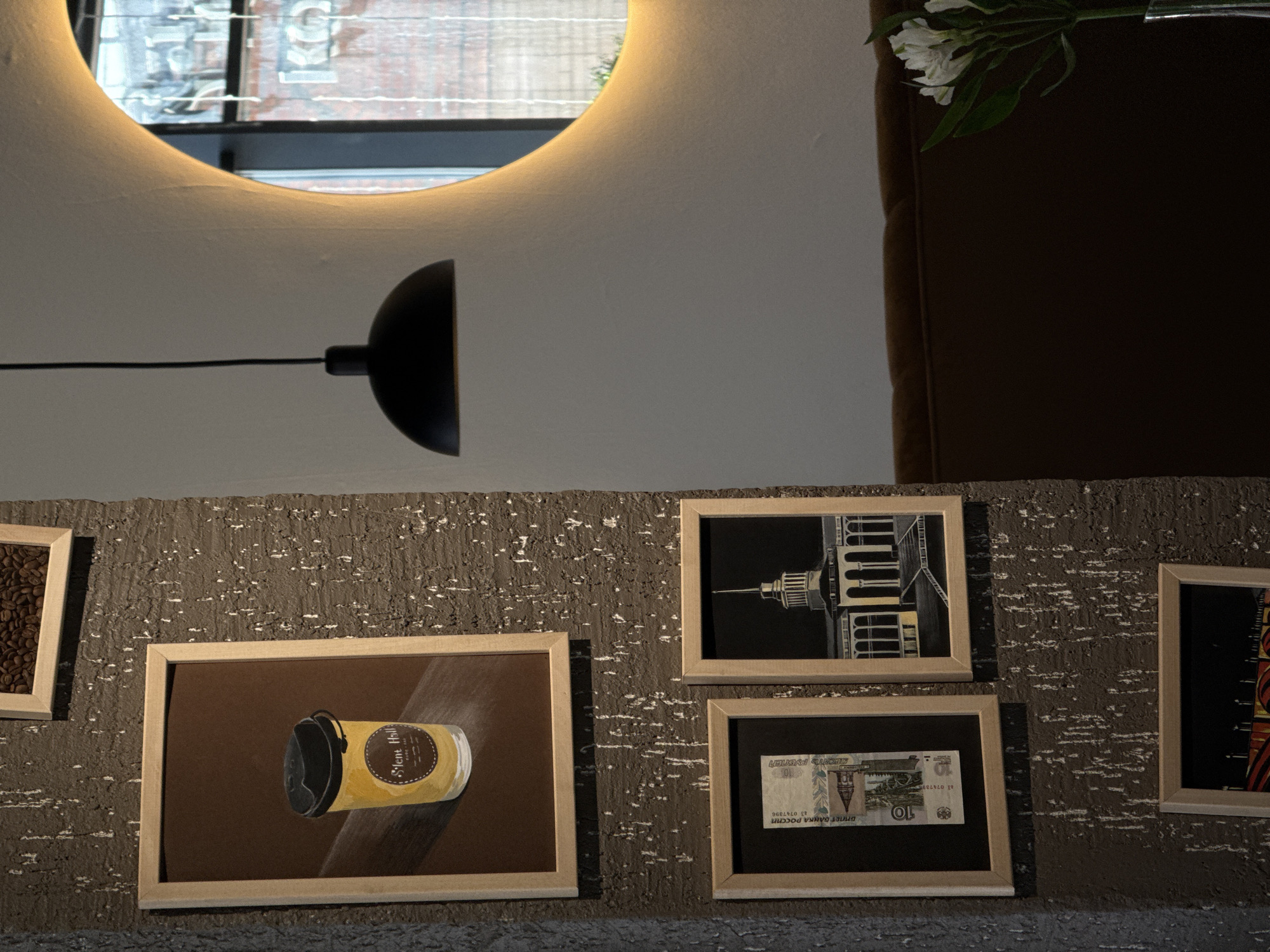 The image shows a wall in an establishment decorated with a variety of framed pictures. One frame displays an image of a coffee cup, another displays coffee beans, another features an architectural drawing of what may be a building, and another features what appears to be Russian currency, a 10 Ruble note. The location may be Krasnoyarsk, Russia. A wall-mounted lamp is visible as well as an upper portion of a mirror. A vase with white flowers is to the upper right. The wall itself is textured.
