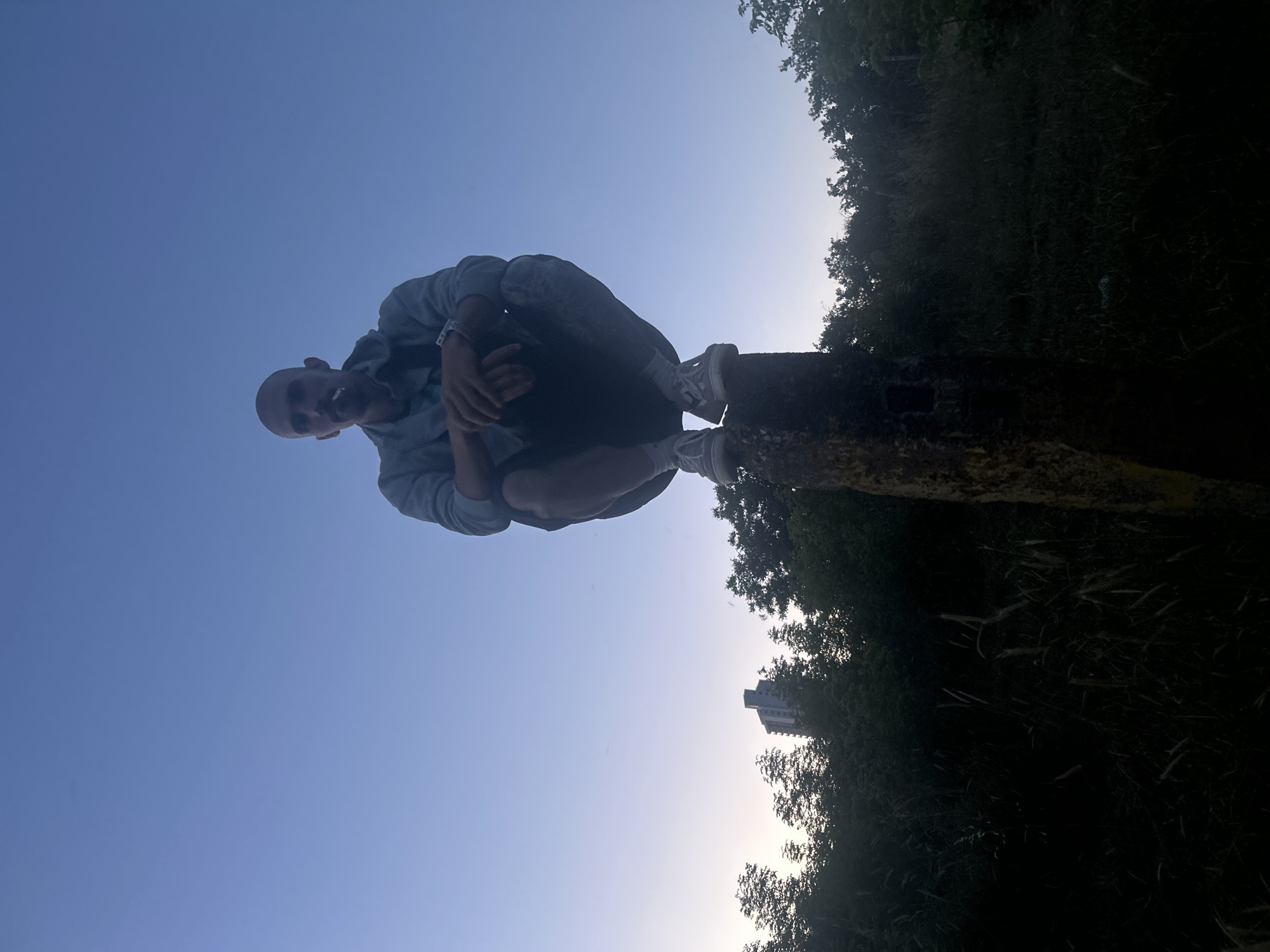 A low-angle, wide shot captures a person squatting on top of a weathered, cylindrical object. The person, who appears to be male, is bald and has a cigarette in their mouth. They are wearing a light-colored hooded jacket over a t-shirt, dark shorts, and white sneakers. Their hands are clasped in front of them. The object they are perched on is roughly waist-high and appears to be made of concrete or stone, with a rough texture and some yellow markings.
The background is dominated by a clear, gradient sky transitioning from a pale blue at the top to a lighter hue near the horizon. Below the sky, a dense line of dark green trees stretches horizontally across the frame. In the distance, peeking above the trees on the left side, is a tall, modern building. The foreground is dark and out of focus, showing a rough texture, possibly grass or foliage, that obscures the ground. The overall lighting suggests either early morning or late evening.
The scene is likely an outdoor, semi-urban or park-like environment. Given the context provided regarding Burgas, Bulgaria, this could be an area on the outskirts of the city, perhaps near a green space or a less developed zone adjacent to urban infrastructure. The specific location within Burgas cannot be determined solely from the visual information present.