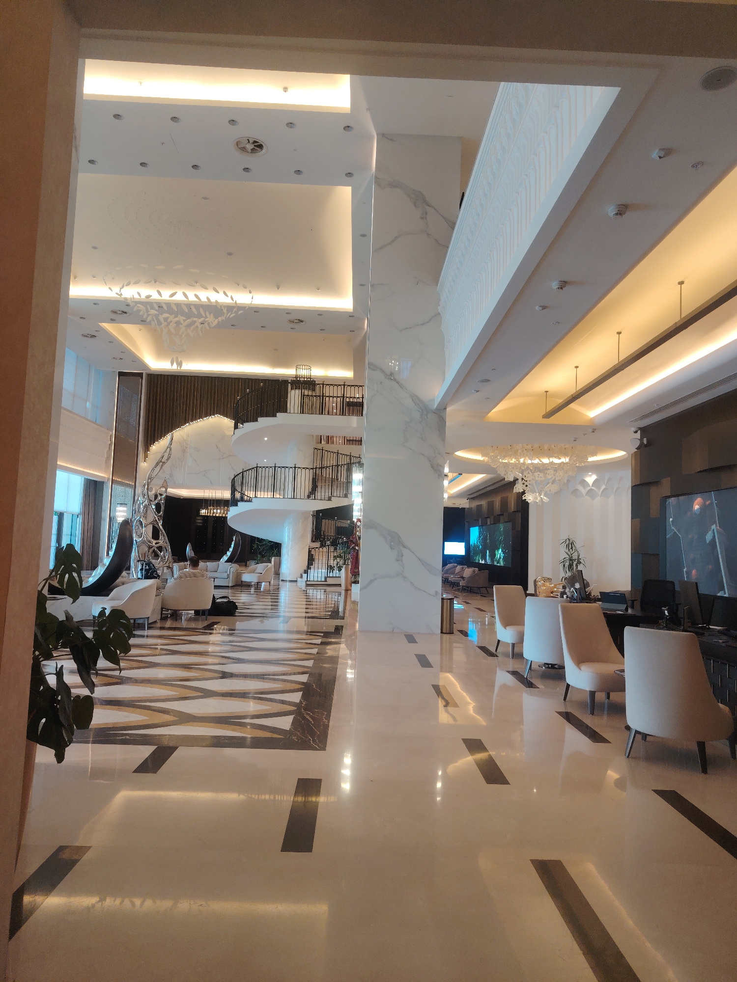 The image depicts the interior of a modern and luxurious hotel lobby, specifically in the city of Konigil, Uzbekistan. The scene is well-lit, with ample natural light coming from large windows and sophisticated artificial lighting systems integrated into the ceiling and walls.
A prominent feature of the lobby is a grand, spiraling staircase with a black metal railing, constructed from white marble. Beside the staircase, a large, abstract metallic sculpture adds a contemporary artistic element. The floor is polished marble with an intricate geometric pattern in shades of cream, brown, and black.
Several seating areas are visible throughout the lobby. In the foreground on the left, there are white upholstered sofas and armchairs arranged around a plant. On the right side of the lobby, a row of light-colored, modern armchairs is positioned, some facing a reception desk which is partially visible.
In the background, large screens are mounted on the walls, displaying video content. The architecture incorporates marble columns with a subtle veining pattern and decorative elements on the upper levels, creating a sense of spaciousness and elegance. The overall impression is one of sophisticated design and comfort.
There are a few individuals present in the scene. One person, wearing a striped shirt, is seated in an armchair in the mid-ground, facing away from the camera, with a black bag on the floor beside them. Other individuals may be present but are not clearly discernible or are out of focus.