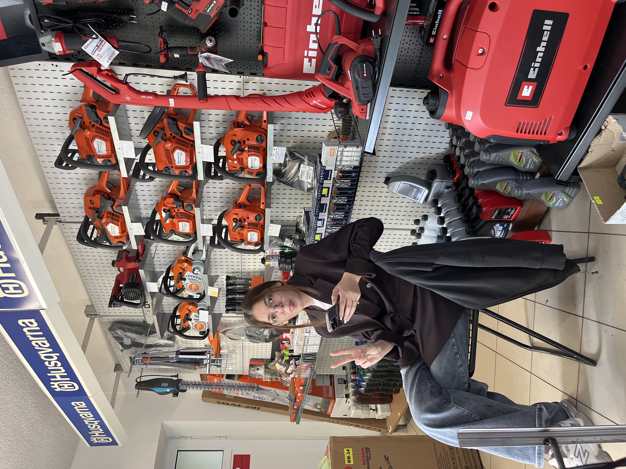 Here's a detailed description of the image:
**Location and Environment:** The image appears to be taken inside a store that sells power tools, possibly in the city of Rudozem, Bulgaria. This is suggested by textual information extracted from the image with OCR. The background shows walls covered with various tools on display, including chainsaws, leaf blowers, and hedge trimmers.
**People:** A young woman is seated on a black metal chair in the middle of the frame. She is wearing glasses, jeans, a brown jacket, and white sneakers. She is holding a cell phone in one hand and making a peace sign with the other.
**Objects:** Besides the various tools mentioned, there are oil bottles, boxes with labels, and a sign displaying "Husqvarna." The woman is holding a black cell phone.
**Action and Interactions:** The woman is posing for a picture, as suggested by her hand gesture and phone positioning.
**Forensic details:** The chainsaws are prominently displayed and are likely a main product of the store. The brands visible are Husqvarna and Einhell. The products appear well-organized and ready for sale.