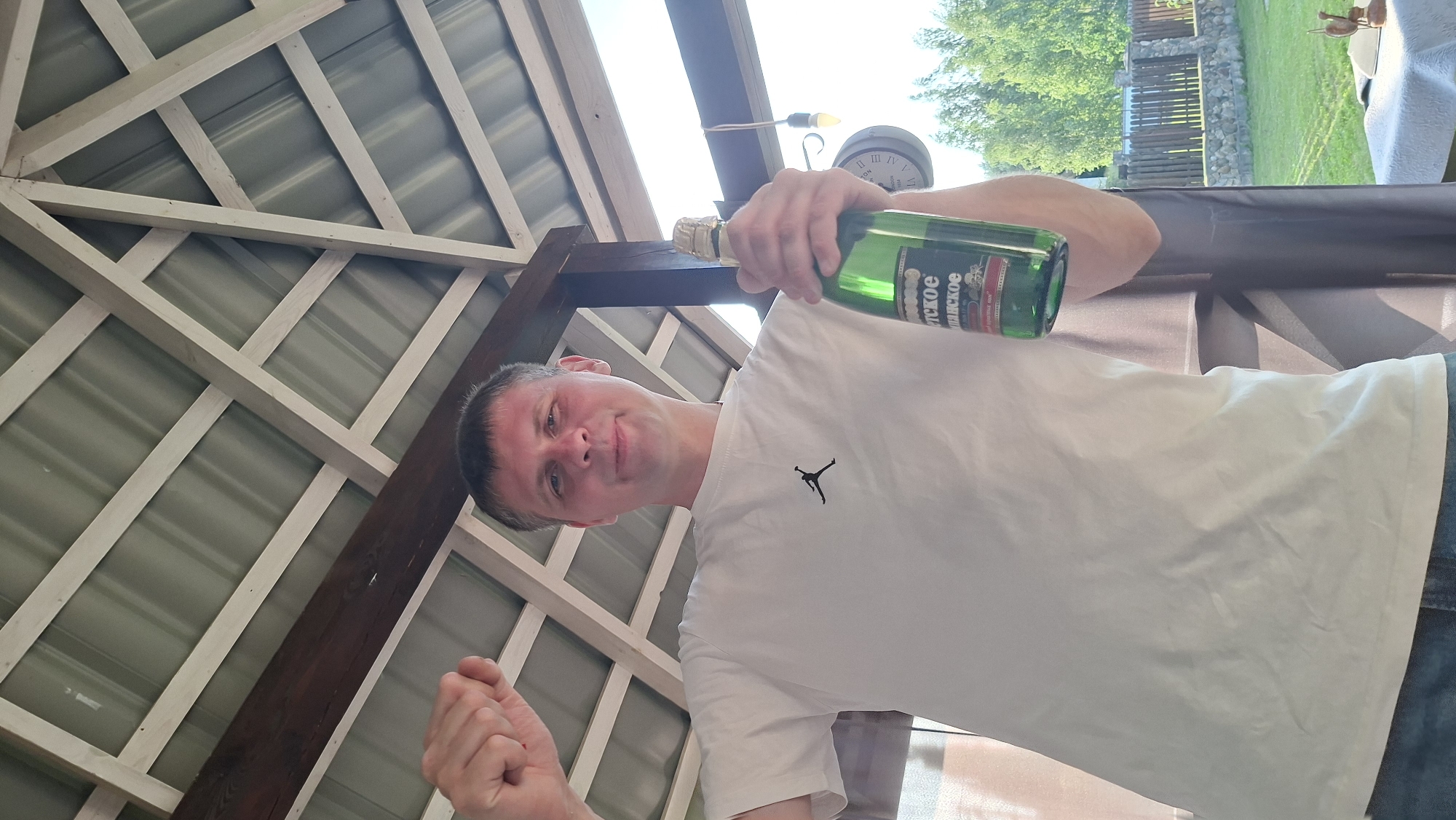 A Caucasian man in his late 30s to early 40s, with short brown hair and a fair complexion, stands outdoors under a covered wooden structure with a corrugated metal roof. He is wearing a white t-shirt with a black Jumpman logo on the left chest and blue jeans. The man is holding a green glass bottle of sparkling wine in his right hand, with the label partially visible. His left arm is raised with a clenched fist. The background shows a fence, trees, and a clear sky, suggesting a rural or suburban setting. A wall clock is visible in the background. The image was taken at a low angle, looking up at the man and the structure. The scene does not contain any specific identifiable landmarks or indications of the city of Myadzyel, Belarus.