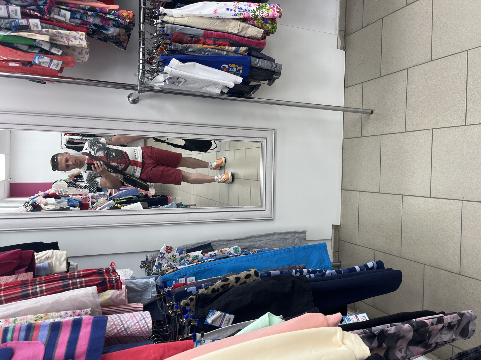 The image depicts the interior of a "Zluka" second-hand clothing store in Minsk, Belarus. An adult male is centered in the frame, captured in his reflection by a large, rectangular, light-framed mirror. He has short, light hair, sunglasses resting on his head, and a short beard. He is wearing a white T-shirt with a dark graphic design, red shorts, and bright orange and white athletic shoes. A dark shoulder bag is slung across his body, and he holds a black smartphone in his right hand, angled towards the mirror, actively taking a picture of his reflection. Tattoos are visible on his left arm.
The environment consists of white walls and light grey rectangular floor tiles with dark grout lines. Clothing racks are positioned on both sides of the mirror and to the right. On the left, multi-tiered racks display various garments, including striped tops, plaid shirts, and pants, with price tags visible, one showing "ЗЛУКА" and "11.90". To the right, a freestanding chrome clothing rack is laden with a diverse assortment of garments, including T-shirts, dresses, and patterned bottoms, some with price tags indicating values like "8.90" and "3.90". A dark bag rests on the floor near the reflected image of the man's feet.