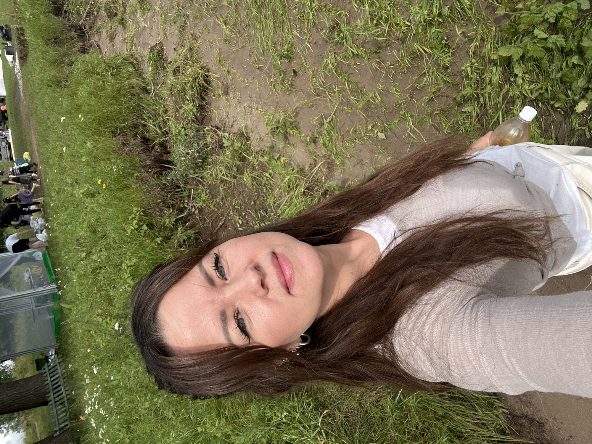 A woman with long brown hair and blue eyes is taking a selfie outdoors. She is wearing a beige long-sleeved shirt and white pants. She is holding a clear plastic bottle with a white cap in her right hand. The background shows a grassy area with some small white and yellow flowers, and in the distance, a greenhouse-like structure and several people. The terrain appears to be uneven with patches of mud and grass. The location is stated to be Chaikovsky, Russia, suggesting a park or outdoor recreational area.