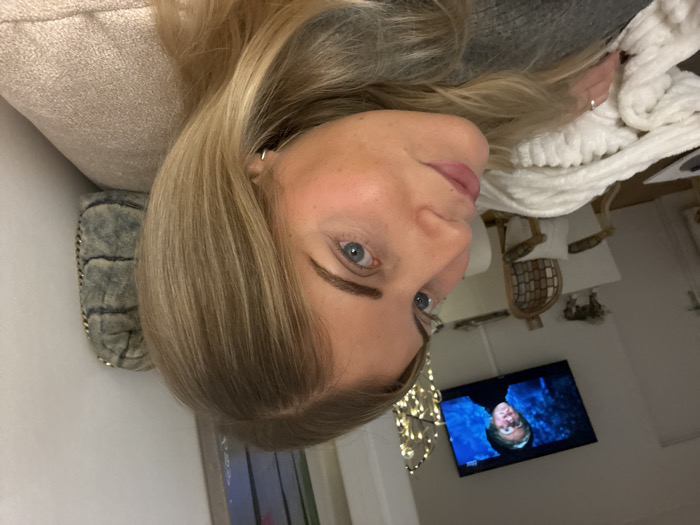 A woman with blonde hair and blue eyes is seen in a portrait. She appears to be lying down, possibly on a couch with a decorative pillow. In the background, there's a TV displaying a scene from what appears to be the movie "Harry Potter." A fluffy white throw blanket and a glimpse of a decorative chair are also visible. The scene suggests a cozy, relaxed atmosphere.