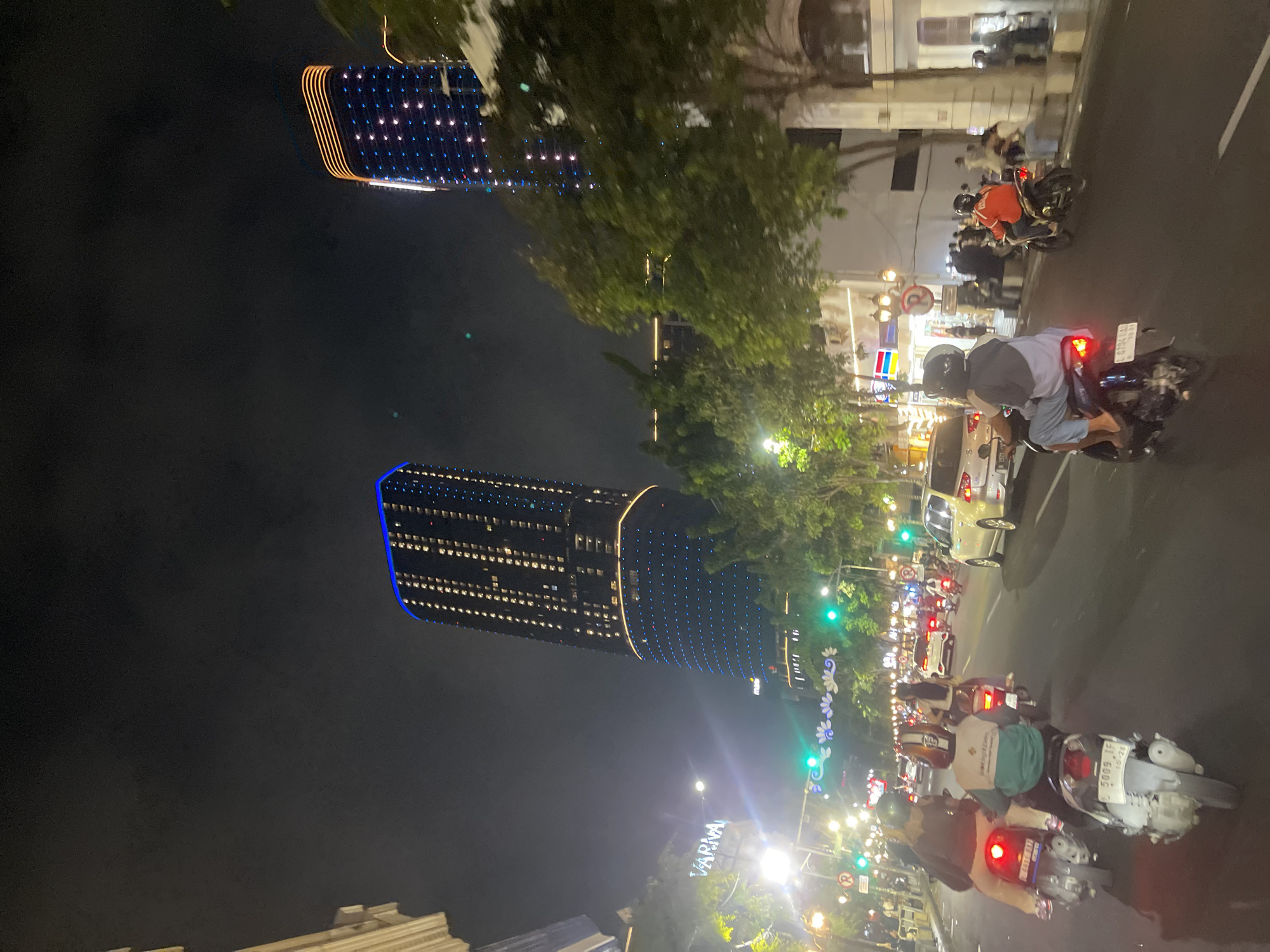 The image depicts a nighttime scene on a city street, likely in Surabaya, Indonesia, given the presence of distinctive skyscrapers and the overall urban ambiance. The foreground is dominated by several motorcycles with riders, navigating a busy road. Traffic lights are visible and appear to be green, indicating the flow of vehicles. On the right side of the street, people are gathered, some seated on a low wall, while others are standing. Trees line the sidewalk, and there are illuminated buildings and signs in the background. The most prominent features are two tall, modern skyscrapers, one with blue and orange lights along its exterior, and the other adorned with numerous white and blue lights, creating a striking visual display against the dark night sky. The overall impression is of a vibrant, bustling urban environment at night.
