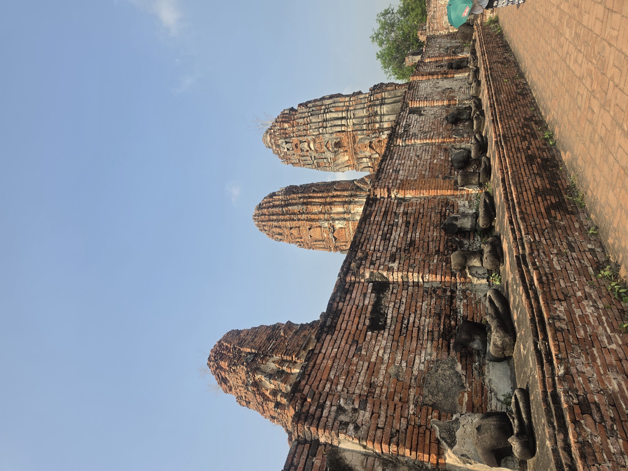 The image captures a view of ancient ruins in Phra Nakhon Si Ayutthaya City Municipality, Thailand. The scene focuses on three brick stupas, partially deteriorated, reaching upwards towards a light blue sky. Adjacent to the stupas, there is a long brick wall, also displaying signs of age and wear. At the base of the wall, remnants of stone carvings, possibly depicting reclining Buddha figures, are visible. Part of a red brick pathway is also visible. A person with a green umbrella is seen on the path.