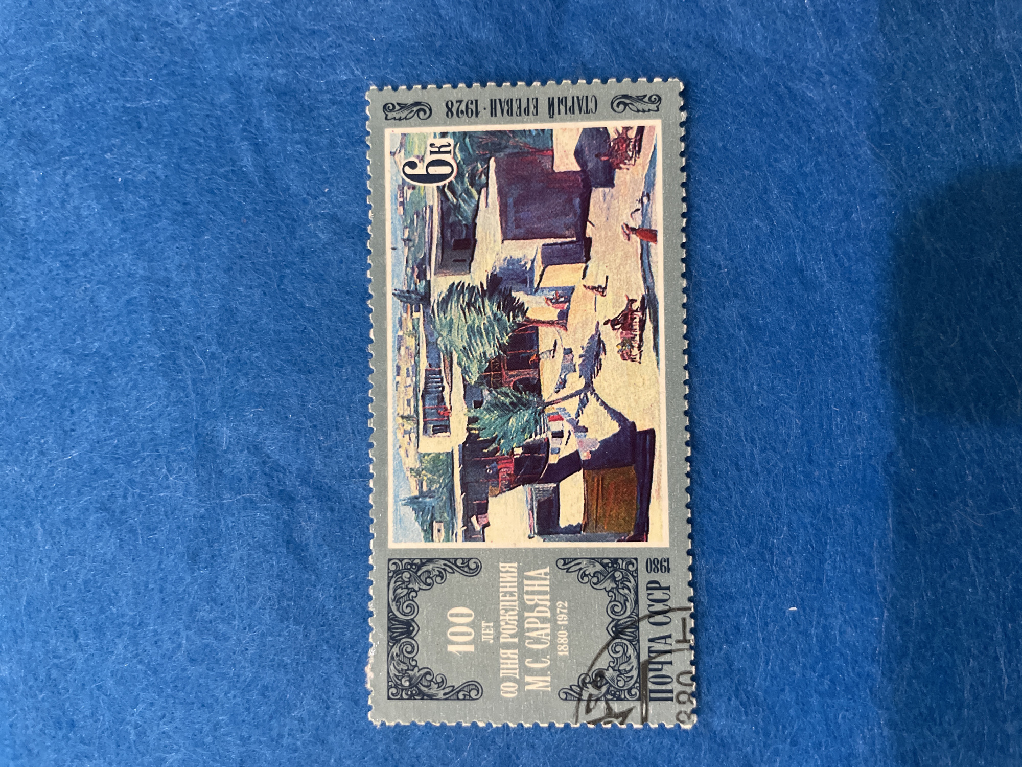 This is a detailed, forensic, matter-of-fact description of a Soviet postage stamp, not an image of the city of Perm, Russia.
The image is a postage stamp with a light blue border and a perforated edge. The stamp features a decorative ornate frame on the left side, enclosing text and numbers in a dark gray color. The text reads "100 ЛЕТ" (100 years) above "СО ДНЯ РОЖДЕНИЯ" (since the birth of) and "М. С. САРЬЯНА" (M. S. Sarian) below that, with "1880-1972" at the bottom of this section. Below this text block is "ПОЧТА СССР" (Post of the USSR) with "1980" vertically to the right and a partial numeral "320" and symbol to the left.
The right side of the stamp depicts a painted scene of an outdoor courtyard. The scene includes low-rise, flat-roofed buildings with simple geometric shapes, suggesting an arid or warm climate. There are sparse trees with windswept foliage, and laundry is hung on a line. Several small figures are depicted in the courtyard. One figure on the right appears to be a woman in a red garment. In the center-left, two figures are seated on a horse-drawn cart. Another figure walks towards the left. In the background, more buildings and a distant landscape are visible under a blue sky. A large "6к" (6 kopecks) is prominently displayed in the upper right corner of the painted scene. Along the right edge of the stamp, vertically oriented text reads "СТАРЫЙ ЕРЕВАН-1928" (Old Yerevan-1928).