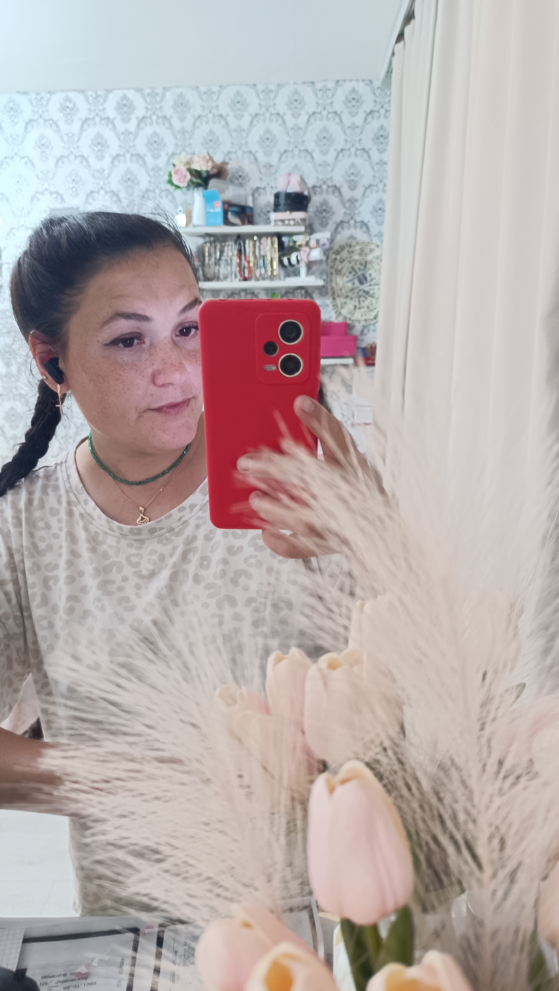 A woman with braided dark hair is taking a selfie in a mirror. She is wearing a light-colored t-shirt with a leopard print pattern, a green beaded necklace, and a gold pendant. She has on wireless earbuds. In the background, there are shelves with various items, including what appears to be makeup and jewelry. A white curtain is visible to the right, and a bouquet of pale pink tulips and white feathery plumes is in the foreground, obscuring part of the lower half of the image. The setting appears to be a domestic indoor space, possibly a bedroom or dressing area, with patterned wallpaper. The image does not provide any specific clues to pinpoint a location within Chaikovsky, Russia.