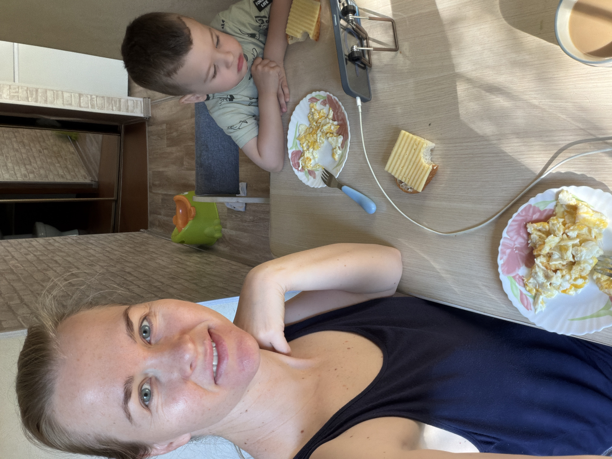 A medium shot captures a woman in a dark blue tank top on the left, and a young boy in a light green t-shirt on the right, both seated at a wooden table. The woman is looking at the camera with a slight smile. The boy is looking down at his phone, which is propped up on a stand. On the table between them are two plates of scrambled eggs, two slices of cheese on toast, a fork, and a mug of tea. The background shows a doorway with mirrored closet doors and a glimpse of a toilet. A small green and orange toy is visible on the floor. The lighting suggests it is daytime, with sunlight casting shadows on the table. The overall scene appears to be a home setting, likely a kitchen or dining area in Khabarovsk, Russia.