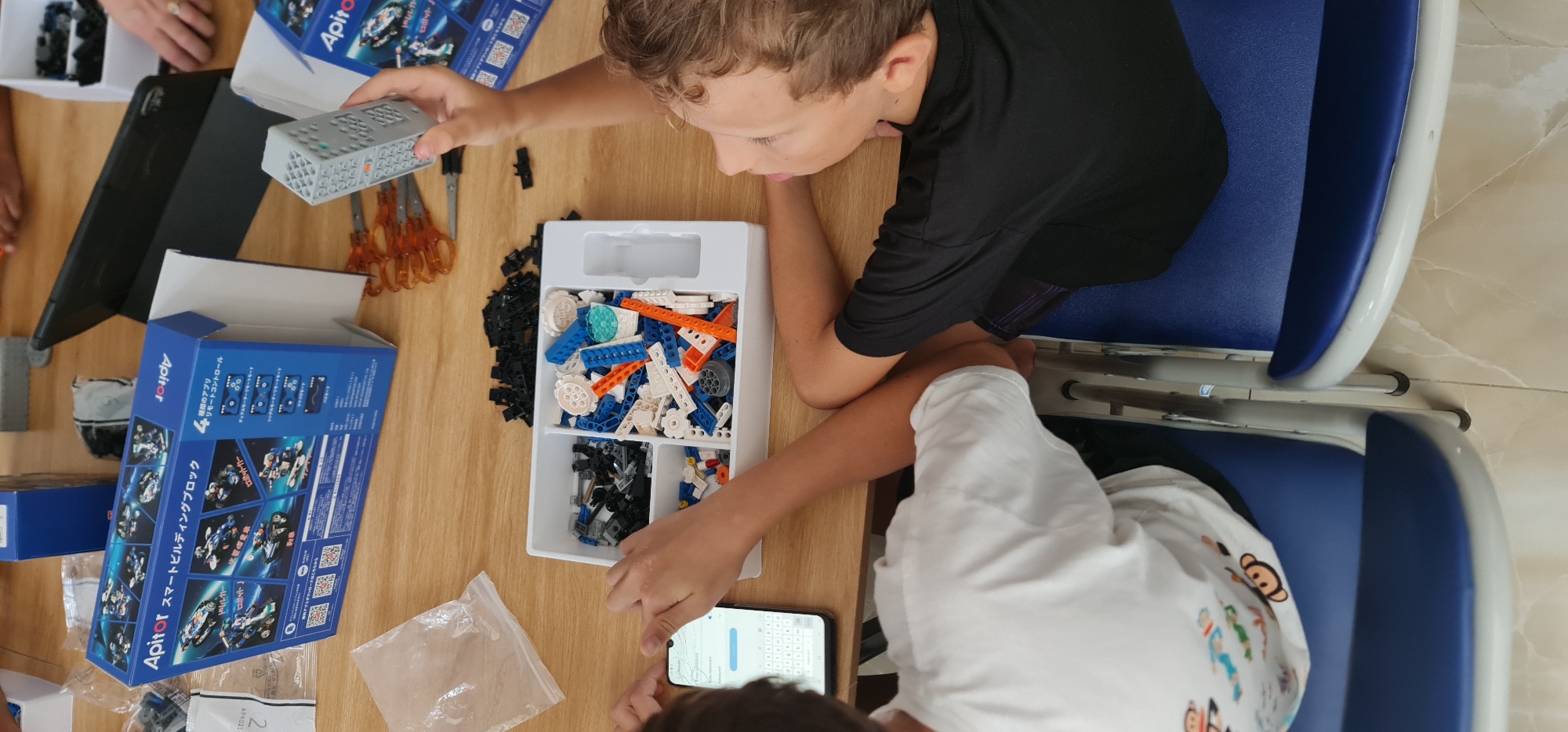 Two children are sitting at a wooden table, assembling an "Apitor Smart Building Block" set. One child, wearing a black shirt, is holding a remote control-like component. The other child, in a white patterned t-shirt, is looking at a smartphone. A box of building blocks, the set's packaging, and various accessories are spread across the table. Chairs with blue and white frames are positioned behind the children. The location is likely indoors. There is no identifiable information indicating this is Đà Nẵng, Vietnam.