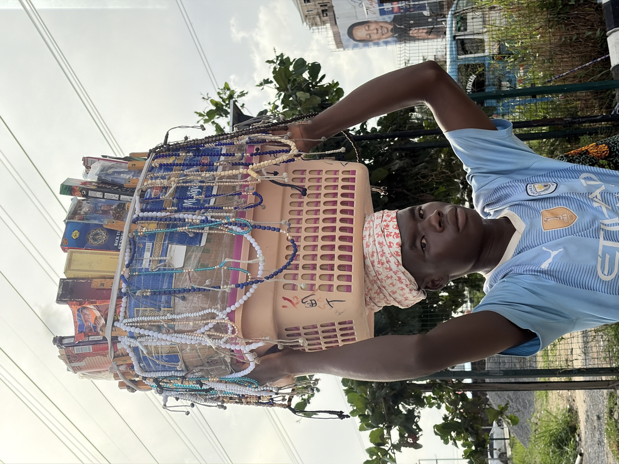 A young Black man, likely a street vendor, is depicted in an outdoor daytime setting in Igboefon, Nigeria. He wears a light blue Manchester City football jersey, complete with the club crest, PUMA logo, "ETIHAD" sponsor, and a FIFA World Champions patch. A white headscarf with red patterns is tied around his head. He looks directly at the camera with a neutral expression.
The man is balancing a large, light-colored (possibly pinkish-beige) plastic basket on his head. The basket is laden with various goods for sale: numerous strings of beads and bracelets in shades of blue, white, beige, brown, silver, and turquoise dangle from its edges. Stacked within and on top of the basket are several boxes, appearing to contain incense sticks or similar items. Visible text on some boxes includes "BACK TO SENDER" and a partial "Mystic" brand name, alongside an image of a person and some illegible script. Red markings, possibly numbers or symbols, are visible on the side of the basket.
The background features an overcast sky with power lines stretching across. Green foliage from trees and bushes is prominent, and a fence or railing can be seen. In the upper right corner, a large billboard displays the portrait of a man. The scene portrays a typical street vending activity in an urban or semi-urban environment under cloudy daylight conditions.