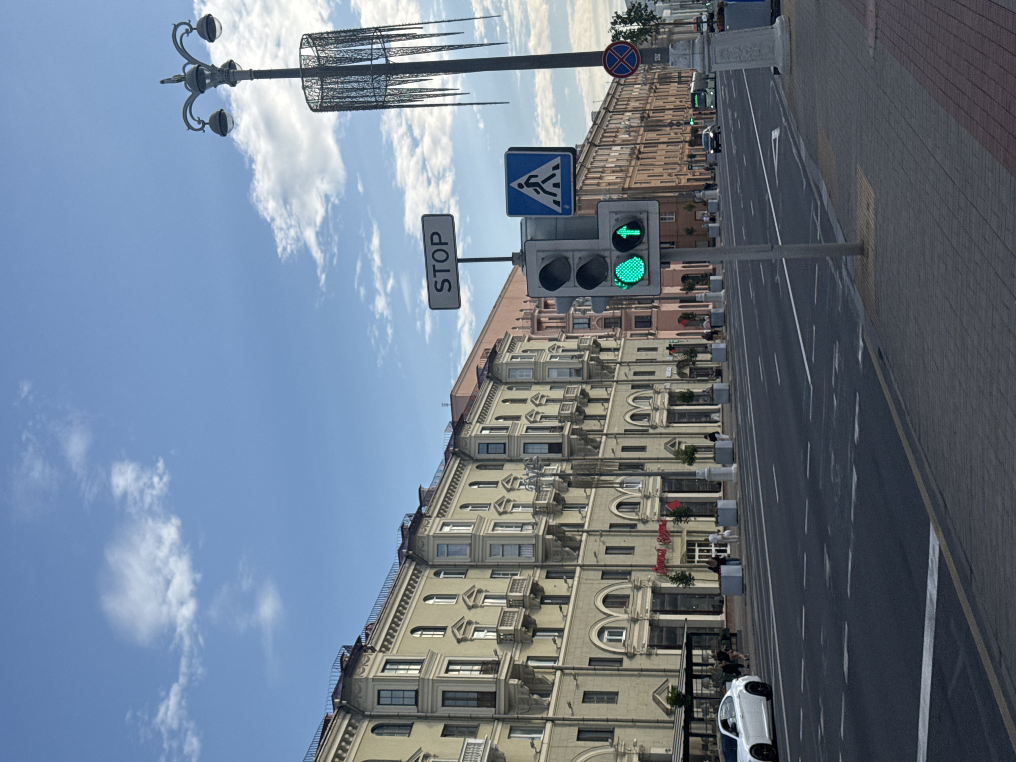 This is a photograph taken from a sidewalk in Minsk, Belarus, looking across a wide, multi-lane street. The street is asphalt with white lane markings and crosswalks. On the left side of the street, there is a row of multi-story buildings with a classical architectural style, featuring balconies, arched windows, and ornate detailing. A white car is parked on the left side of the street, facing left. On the right side of the street, there are more buildings of similar architectural style, with shops and cafes at ground level. Several people are visible walking on the sidewalks and crossing the street. A traffic light is prominently displayed in the foreground, showing a green light for straight traffic and a green arrow for left turns. A pedestrian crossing sign is also visible. Further down the street, traffic can be seen, including cars and a bus. The sky is blue with scattered white clouds. A decorative street lamp with multiple globes stands on the right side of the image. The overall scene depicts a typical urban environment in Minsk.