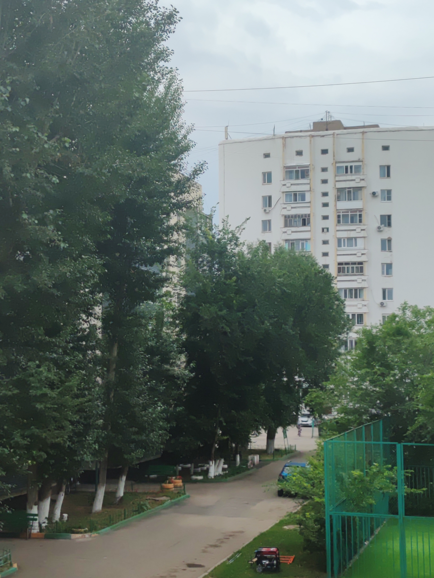 This is an eye-level medium shot taken on an overcast day in Astana, Kazakhstan. The scene depicts an urban environment with a tall, white apartment building dominating the background. The building has multiple floors with numerous windows and air conditioning units visible. In front of the building, a row of tall trees with dense green foliage lines a paved pathway.
The pathway curves to the right, leading away from the viewer and towards the apartment building. Several cars are parked along the side of the pathway, and a small generator with a red and black body and orange attachments is visible on the grass to the right of the pathway. A green metal fence surrounds a green area, possibly a sports court, in the foreground on the right. The trees in the foreground are a prominent feature, their lush leaves creating a natural barrier that partially obscures the view of the apartment building.
There are no people clearly visible in the image, and no text is discernible. The cloudy sky suggests it is daytime, possibly late morning or afternoon, and the overall atmosphere is quiet and suburban, with a focus on the juxtaposition of residential architecture and natural elements. The weather appears to be mild, with no signs of extreme conditions.